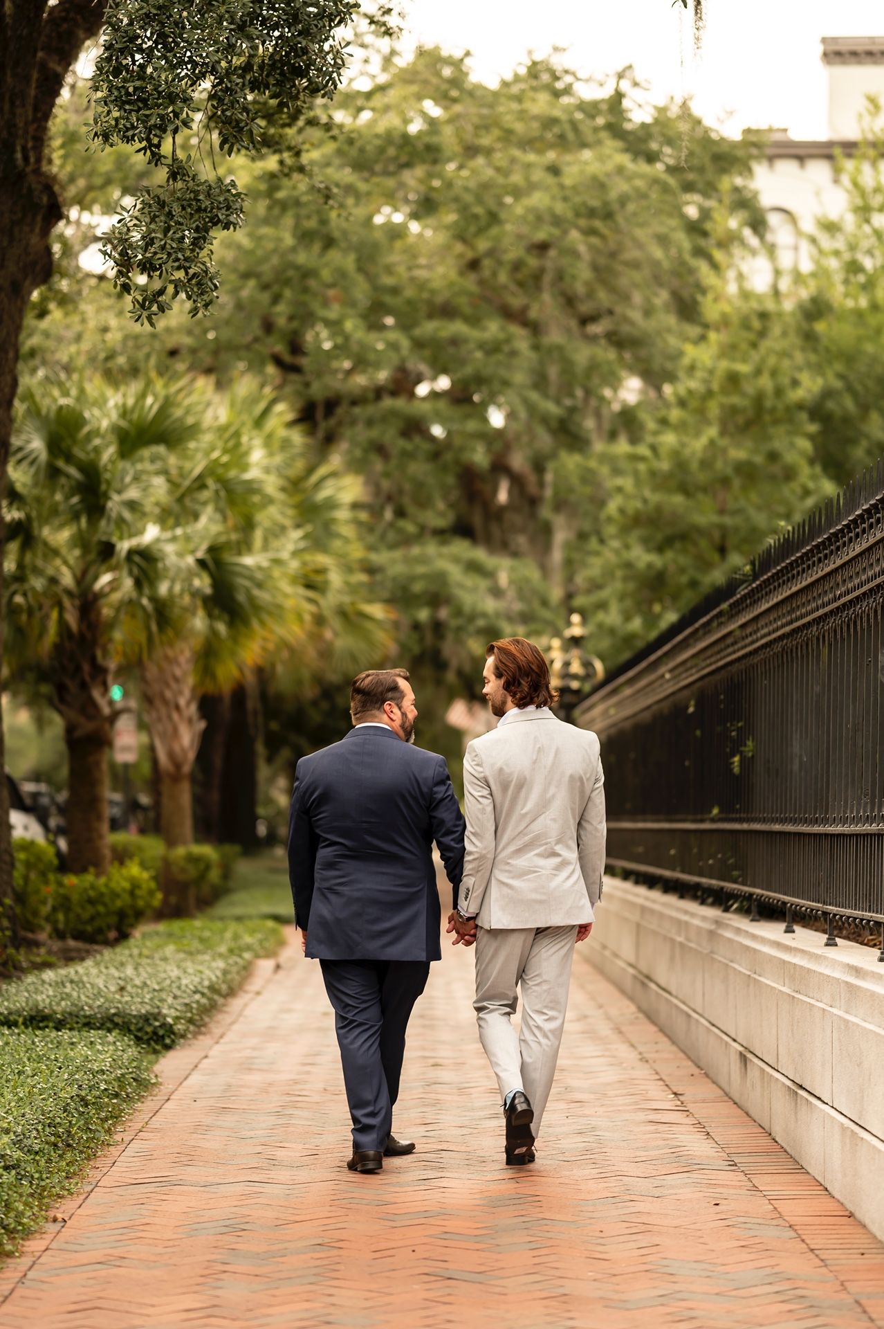 A couple walks hand in hand down a brick pathway surrounded by palm trees and greenery in a garden setting.