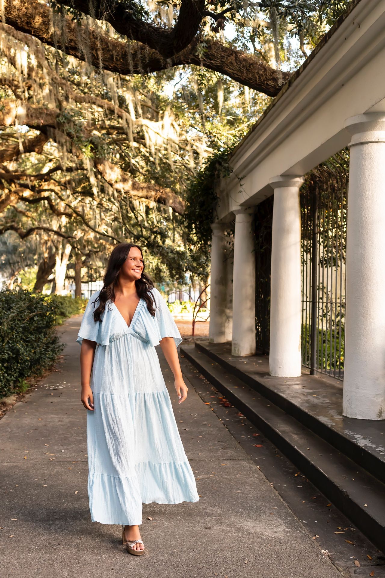 Woman in light blue dress walks along covered walkway with Spanish moss draped oak trees in background.