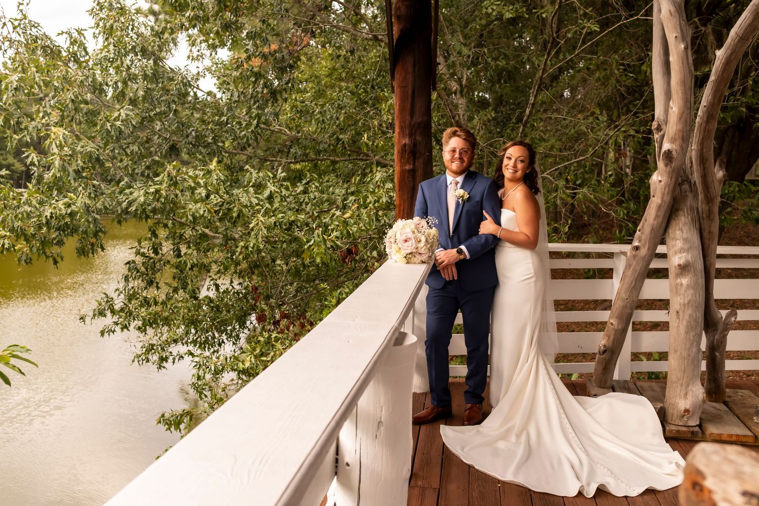 A newlywed couple poses together on a wooden deck overlooking a scenic waterfront view.