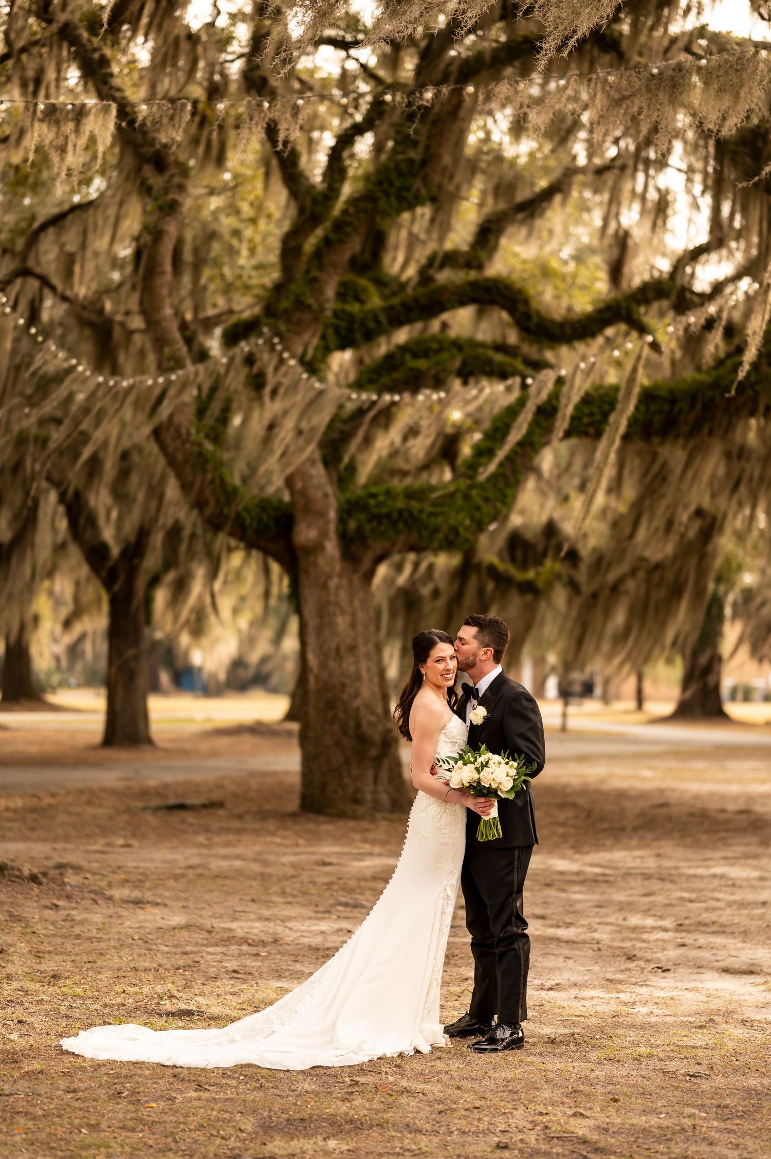 Elegant wedding couple embraces under Spanish moss draped oak trees in romantic outdoor setting.