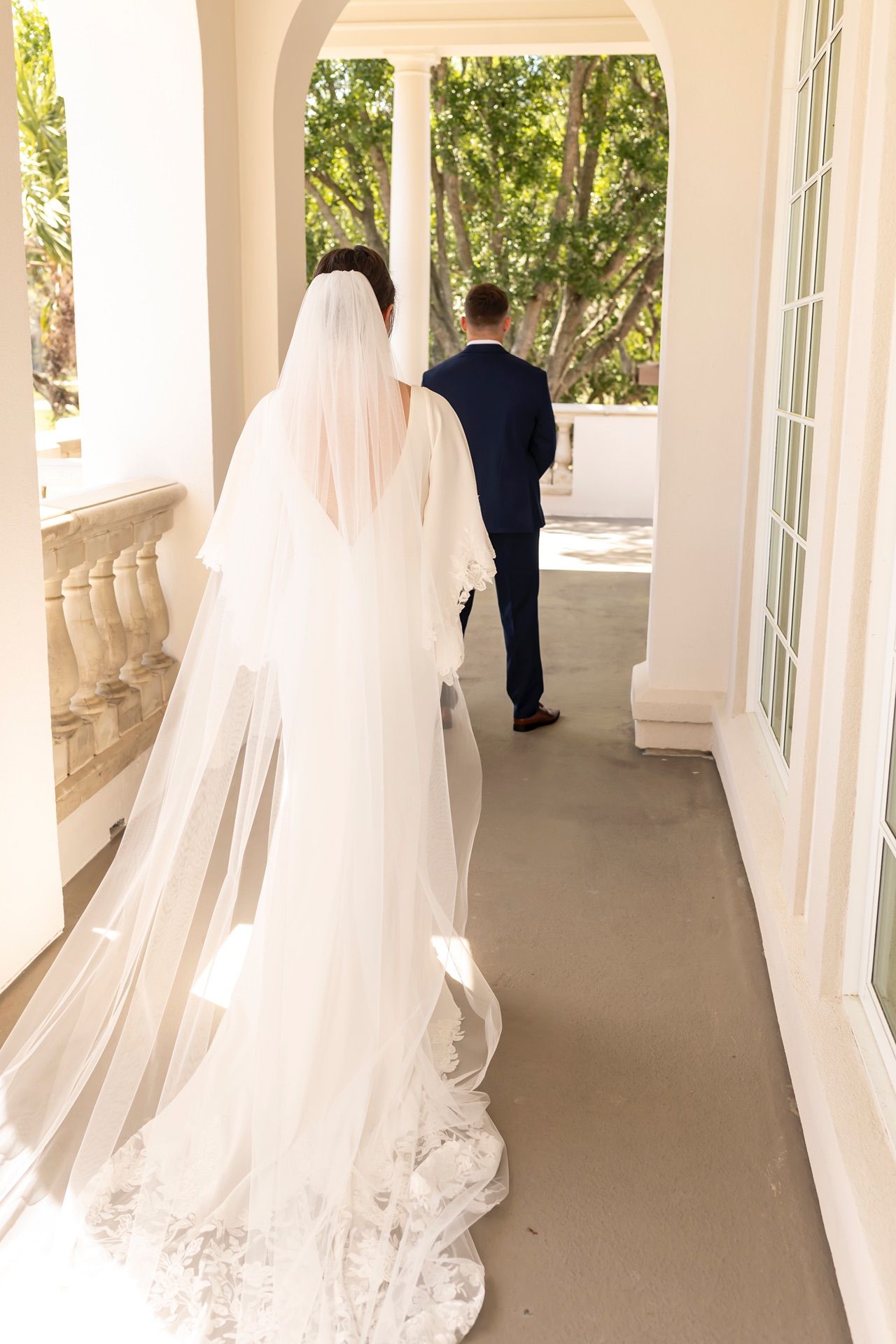A bride in a flowing white veil and dress follows her groom along an elegant covered porch with white columns.