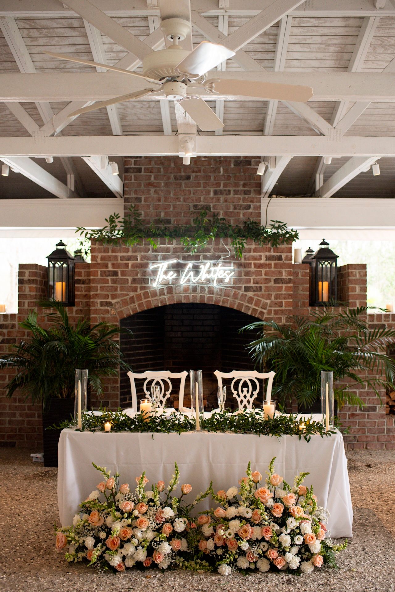 Elegant wedding sweetheart table with peach floral arrangements, greenery, and neon sign above brick fireplace.