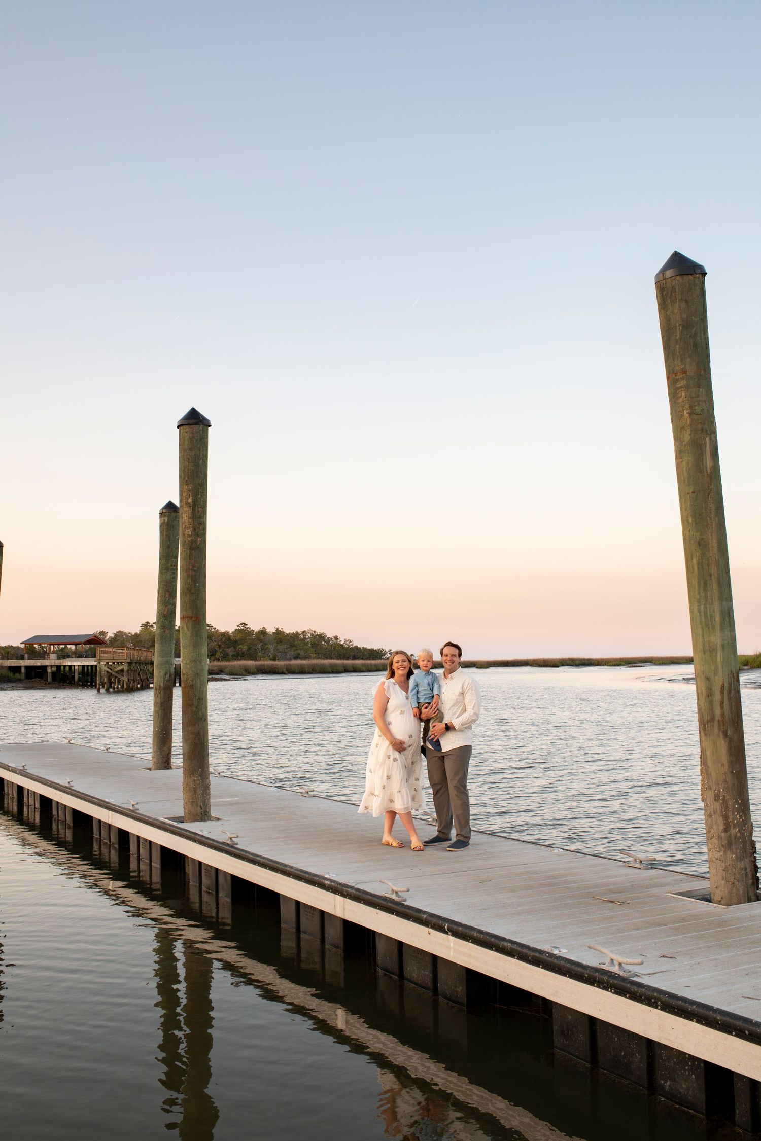 Family of three posing on a wooden dock at sunset with calm water and marsh views in the background.