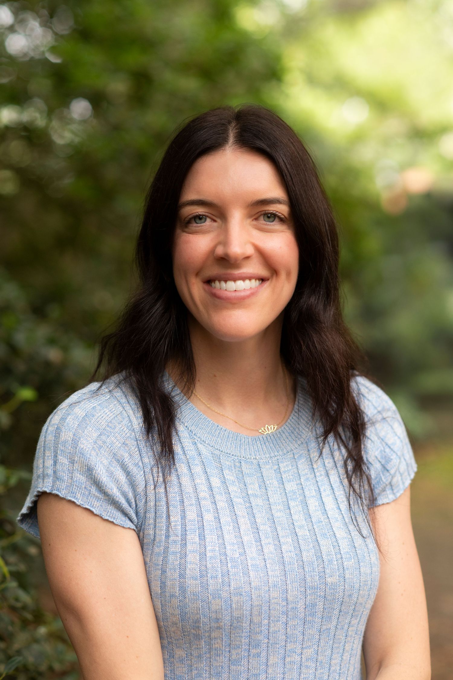 Woman with long dark hair wearing a light blue ribbed top poses outdoors with a bright smile against natural foliage.