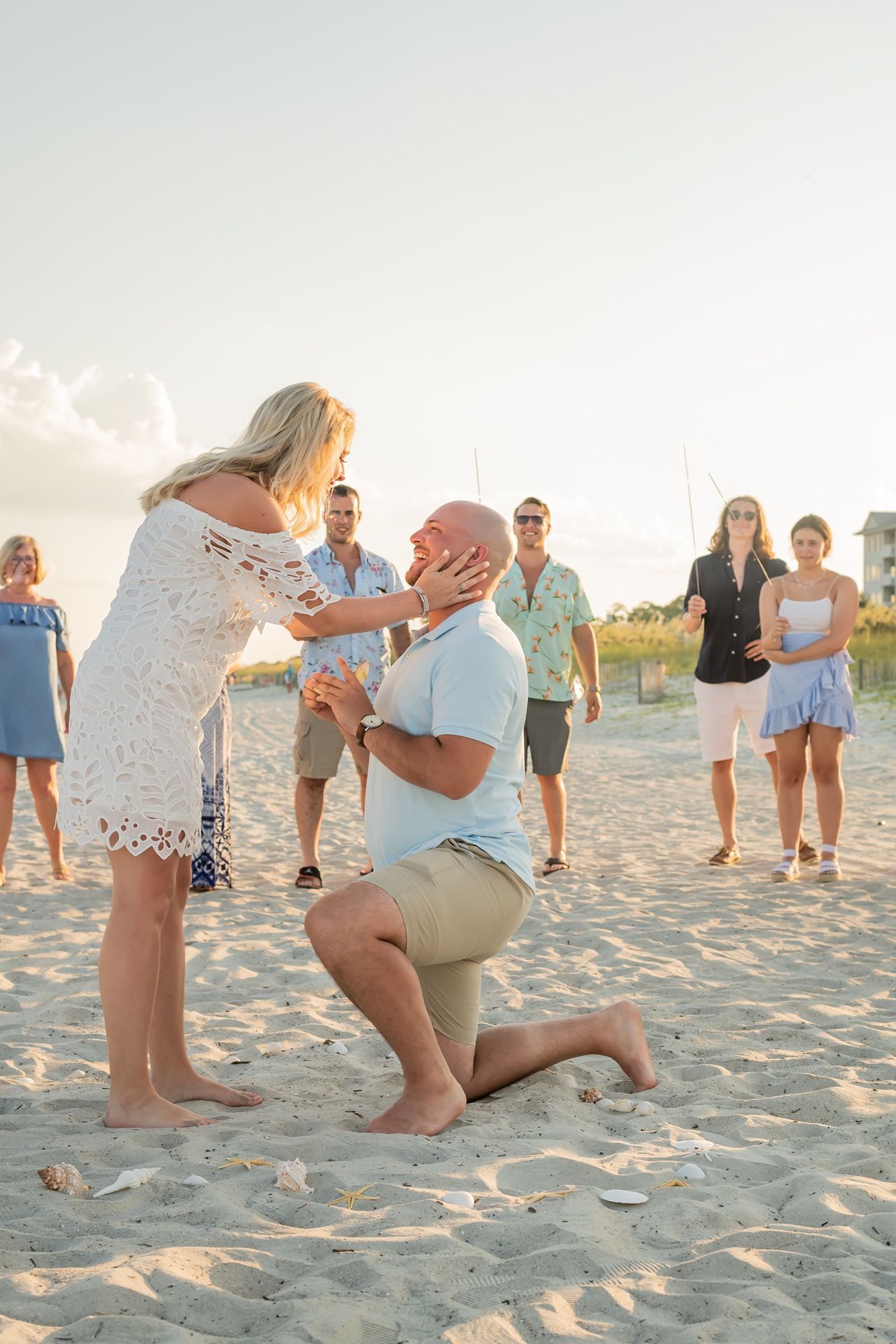 A romantic proposal scene takes place on a sandy beach at sunset with onlookers watching in the background.