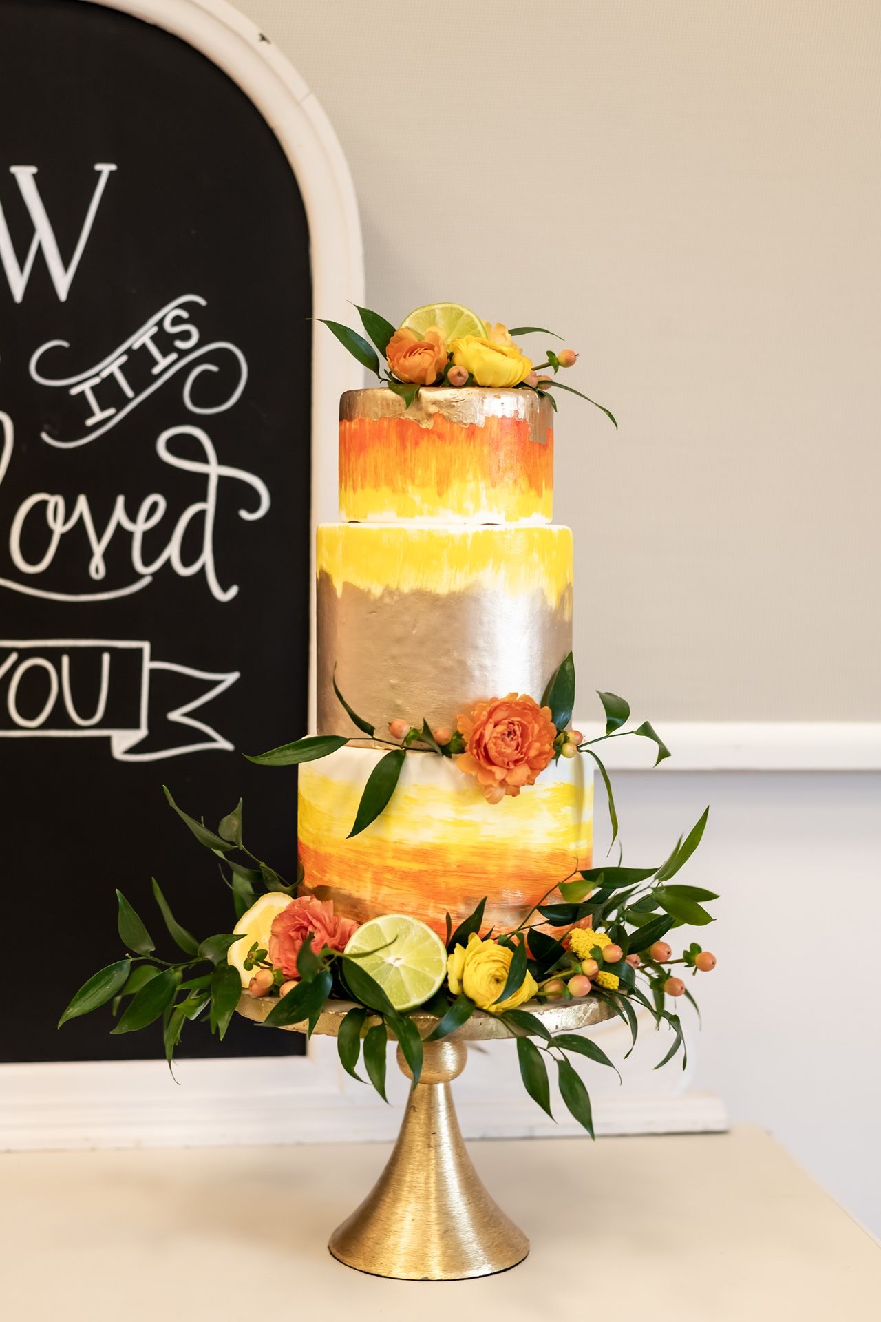 Elegant three-tiered wedding cake with orange watercolor effect and fresh floral decorations on gold metallic stand.
