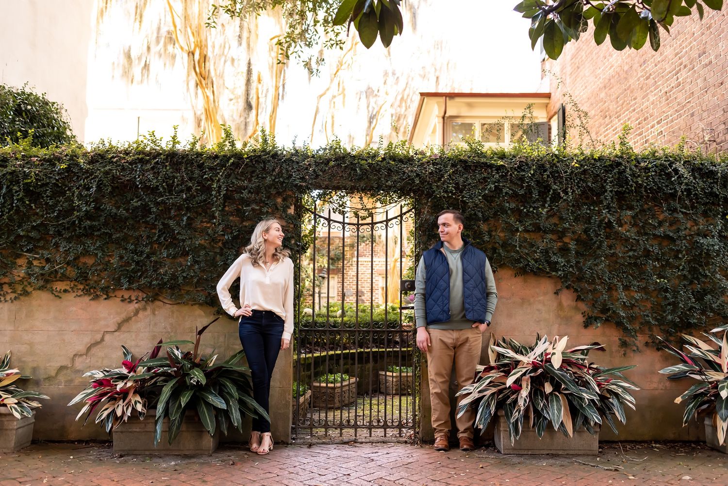 Two people stand beside an ornate iron gate with lush greenery and a pink wall in a courtyard garden setting.