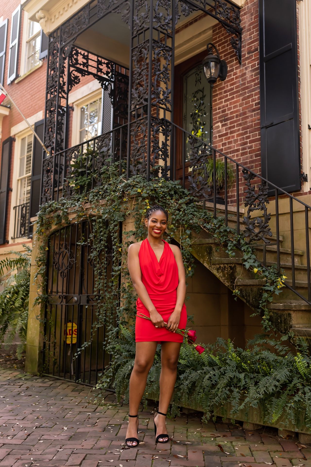 A bright red dress stands out against a historic brick townhouse entrance with ornate iron details and climbing vines.