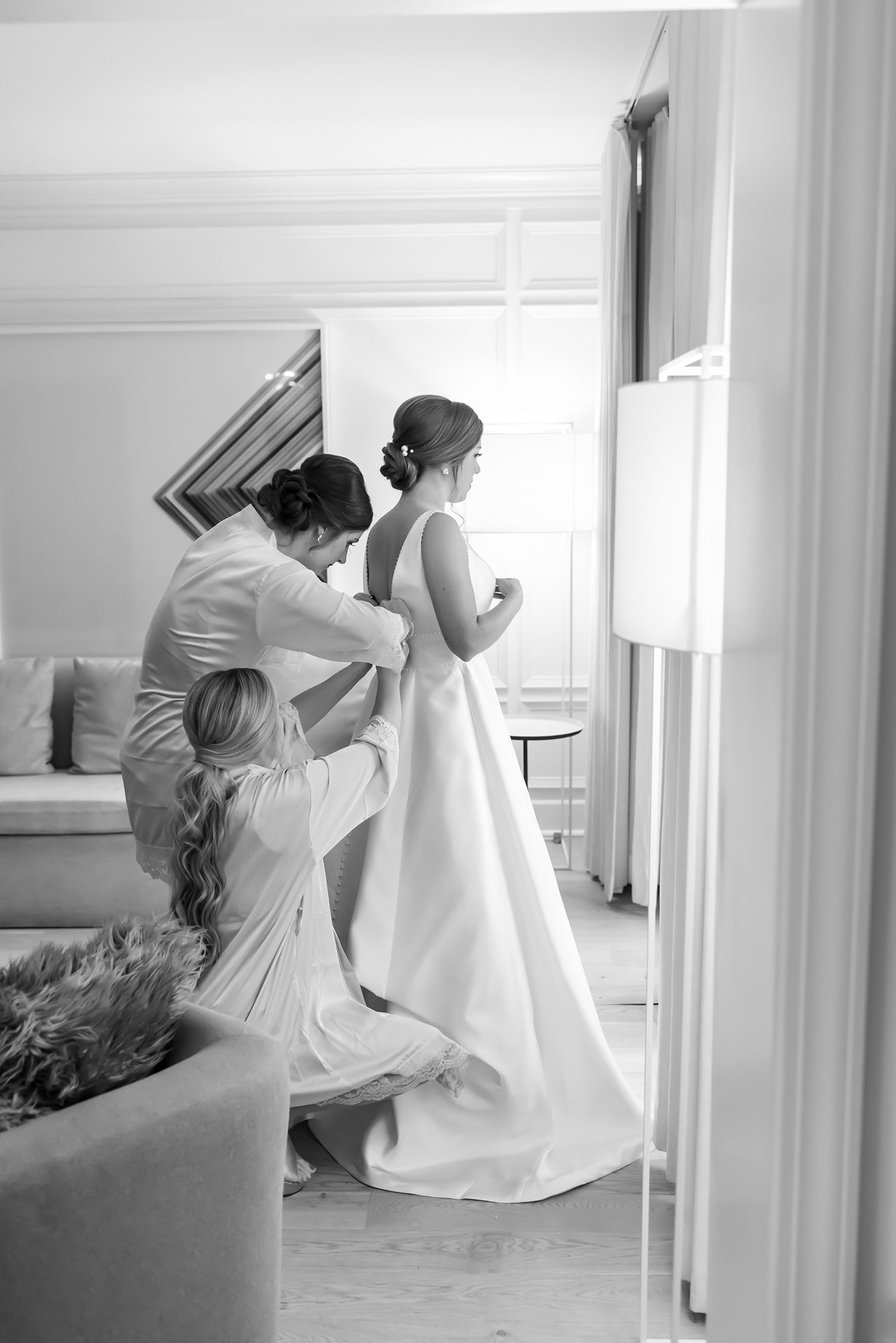 A black and white wedding preparation scene shows final dress adjustments being made in an elegant room.