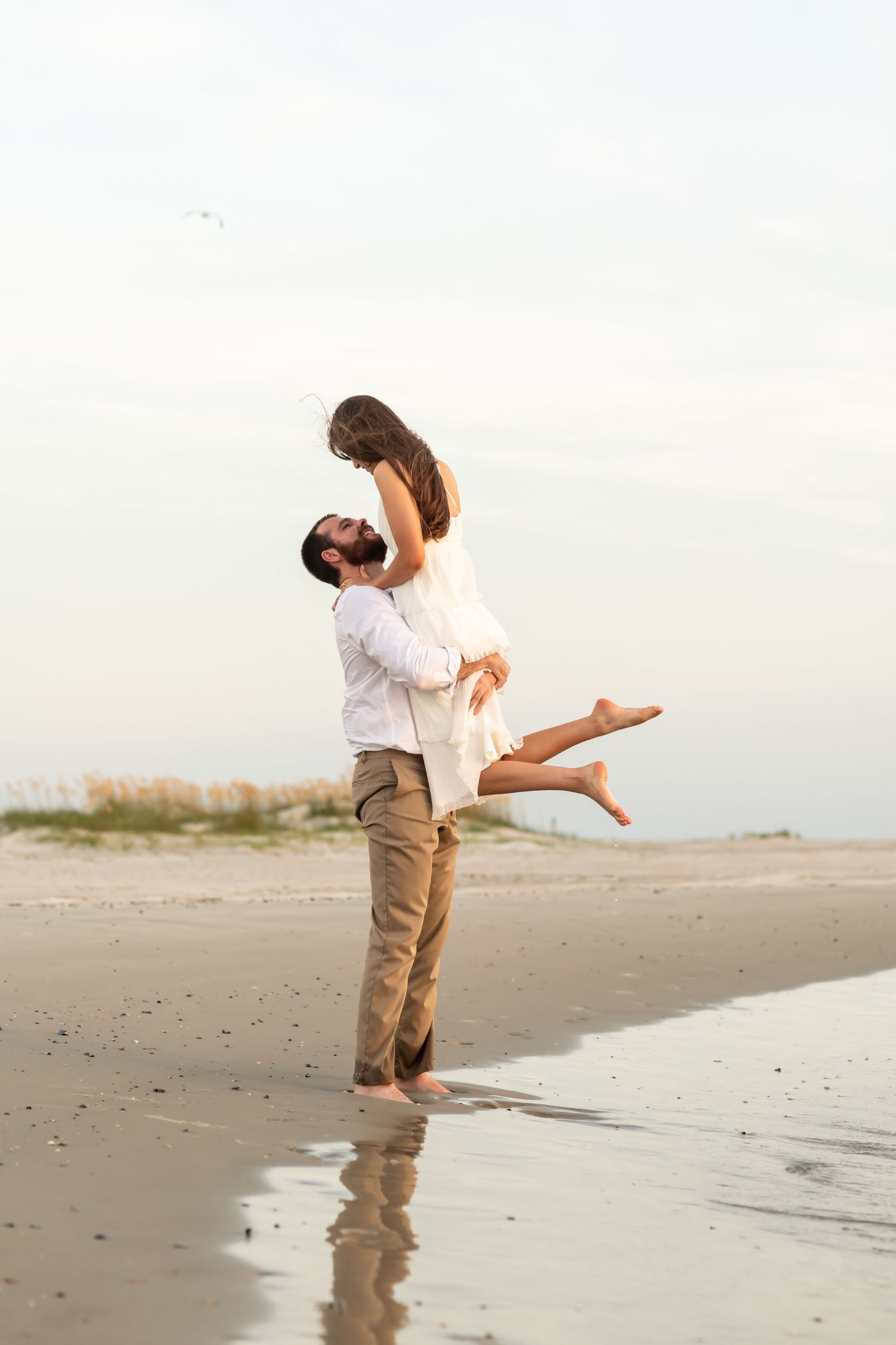 A couple embraces on a beach at sunset, with one person lifted in the air while waves reflect their silhouette.