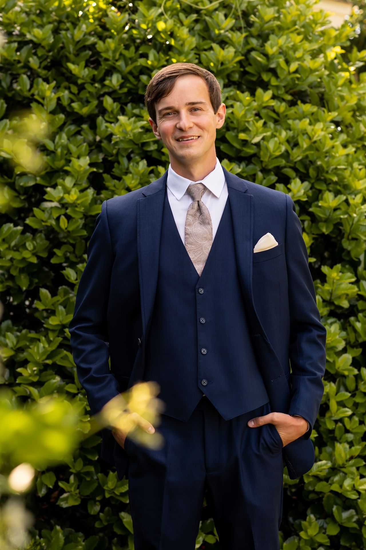 Professional portrait of someone wearing a navy three-piece suit with light tie against green foliage background.