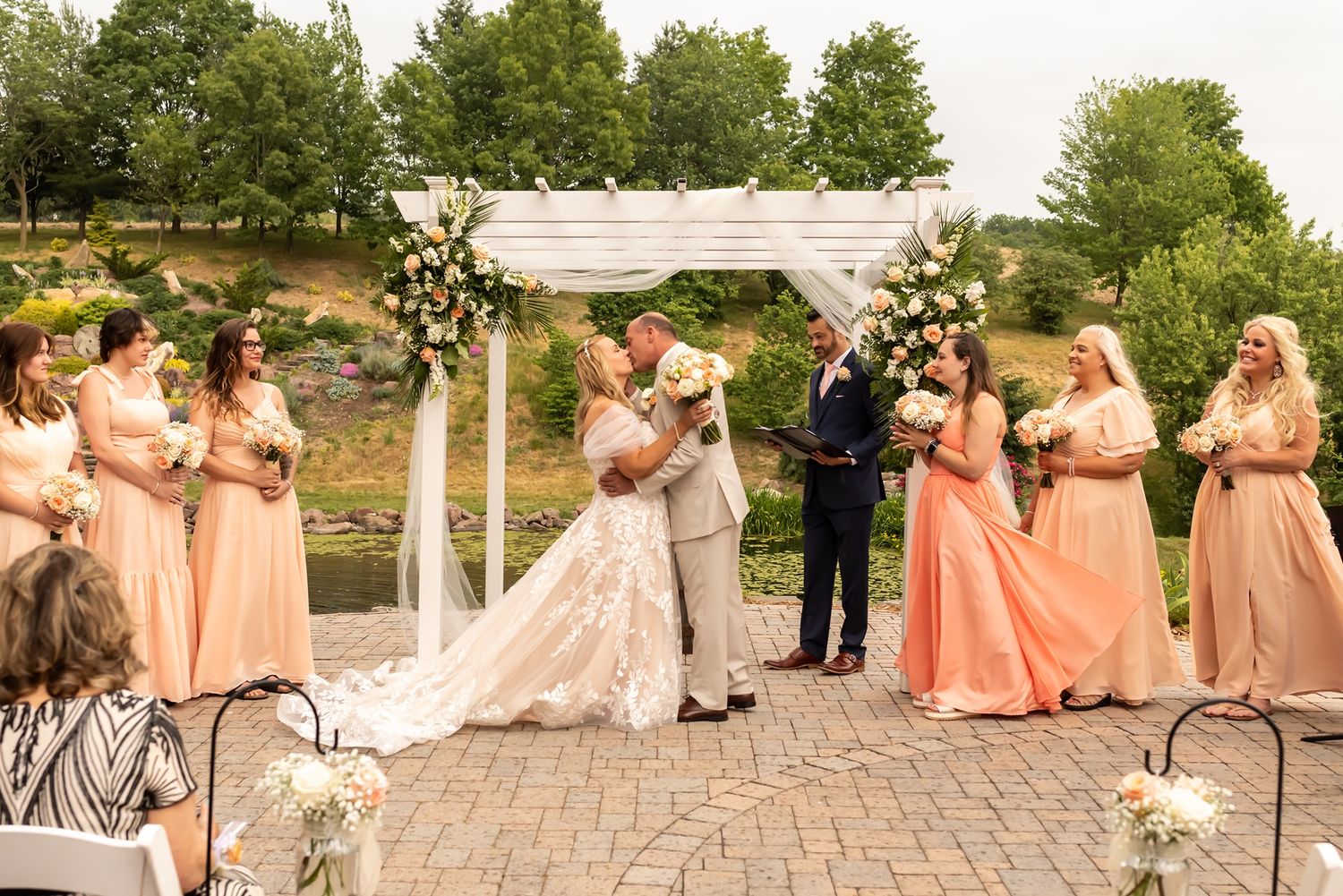 Wedding ceremony takes place outdoors under white arch with bridal party in peach dresses standing on brick patio.