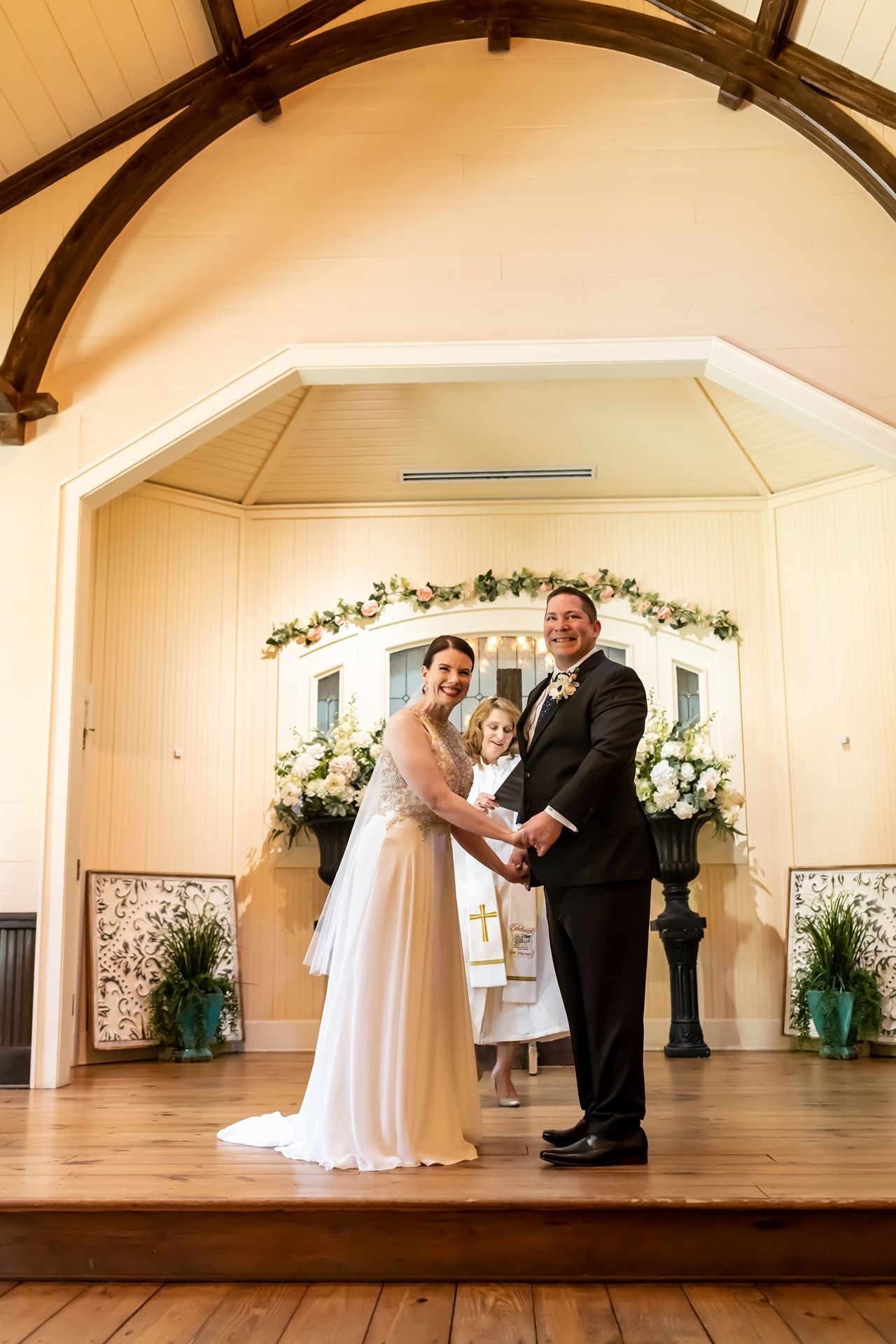 A wedding ceremony takes place on a wooden stage with floral decorations and exposed wooden beams overhead.