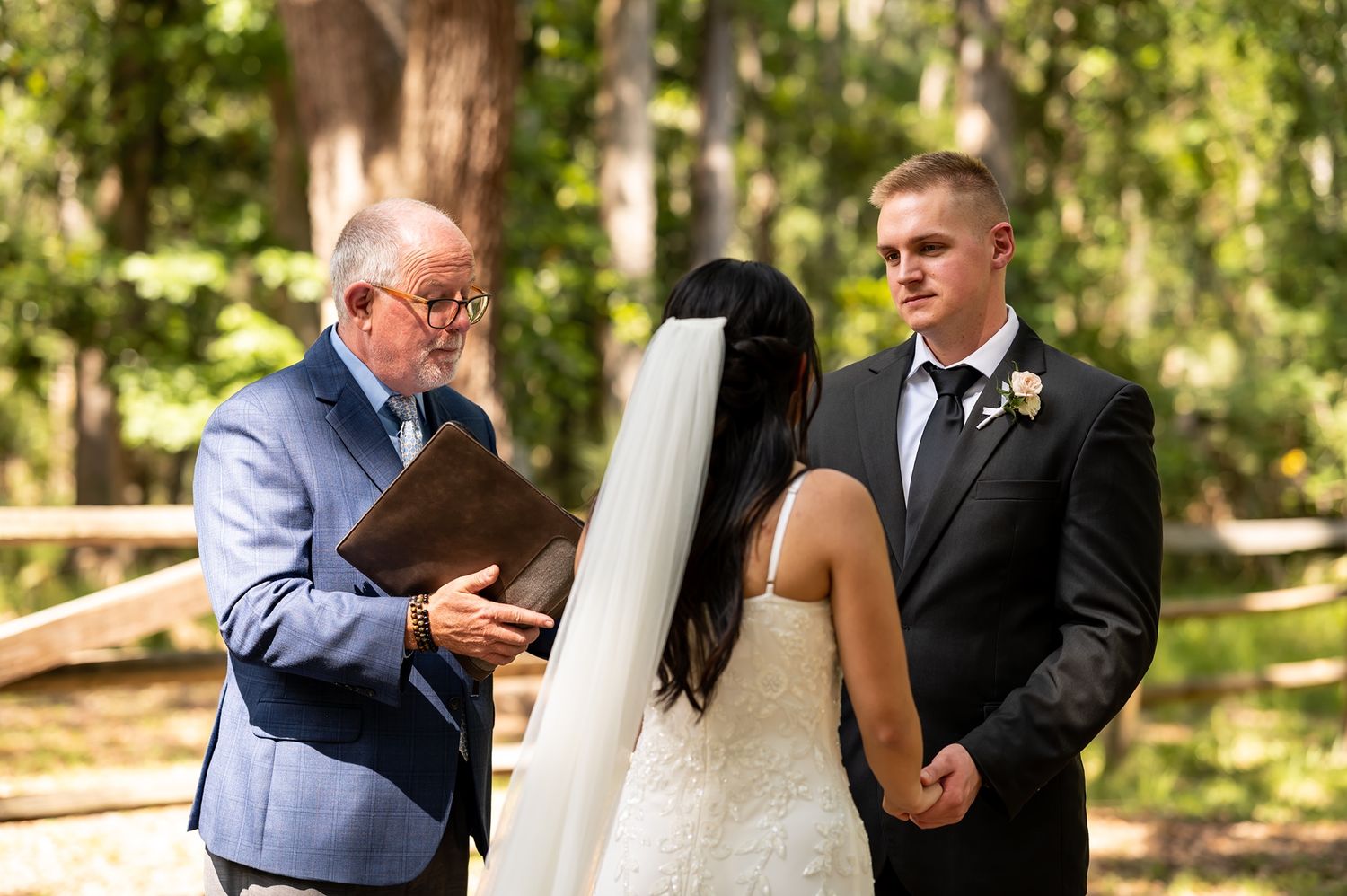 An officiant reads from a book during an outdoor wedding ceremony in a sunlit wooded setting.