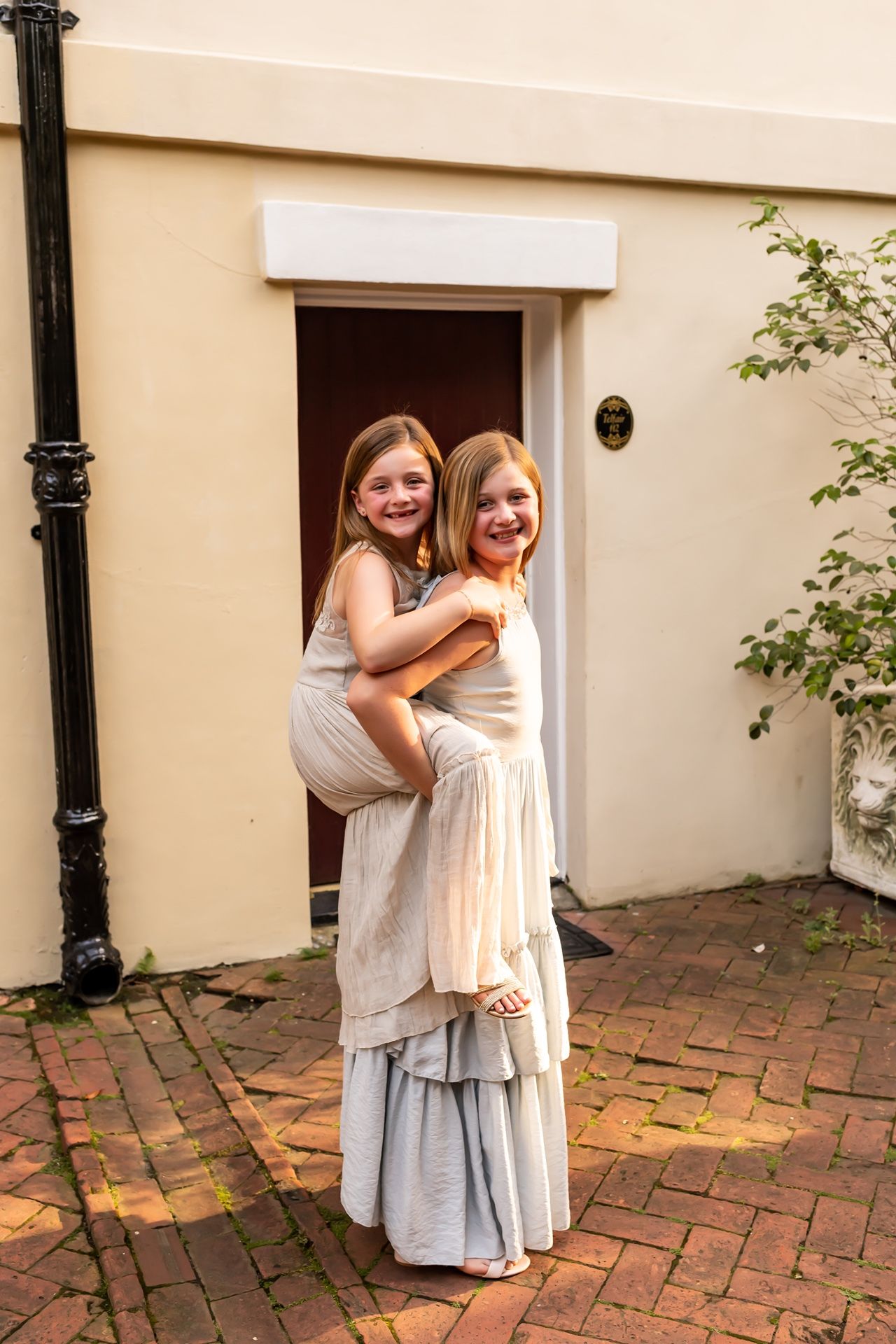 Two young children in light colored dresses hug and play on a red brick patio near a cream colored building.