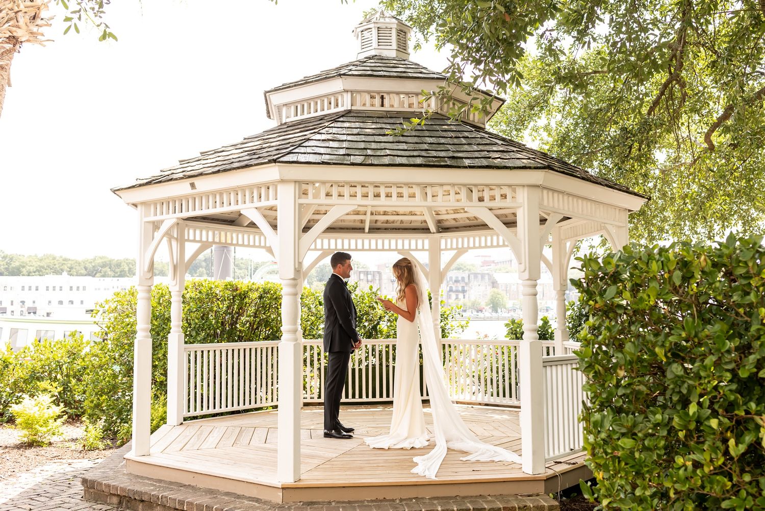 White wooden waterfront gazebo surrounded by lush greenery with wedding couple embraced in coastal afternoon light.