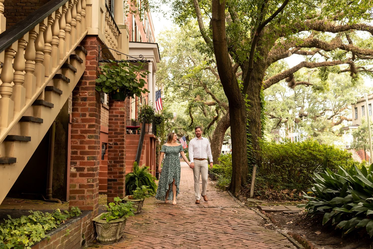 Two people stroll along a charming brick sidewalk under oak trees in historic Savannah's residential district.