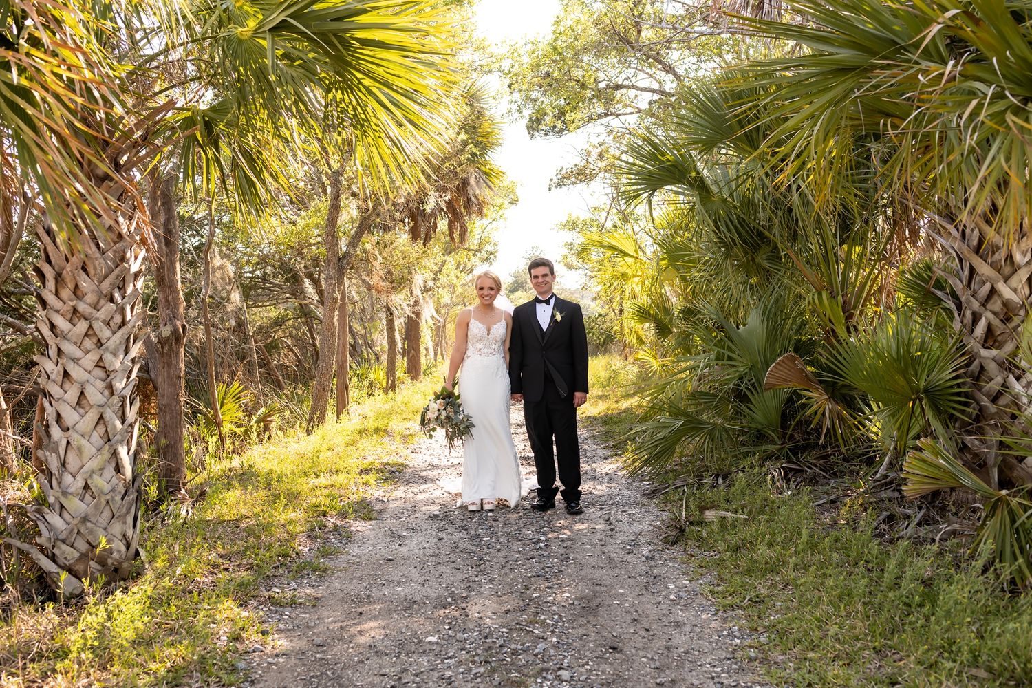 A couple walking down a sandy palm-lined path through natural Florida vegetation on a sunny afternoon.