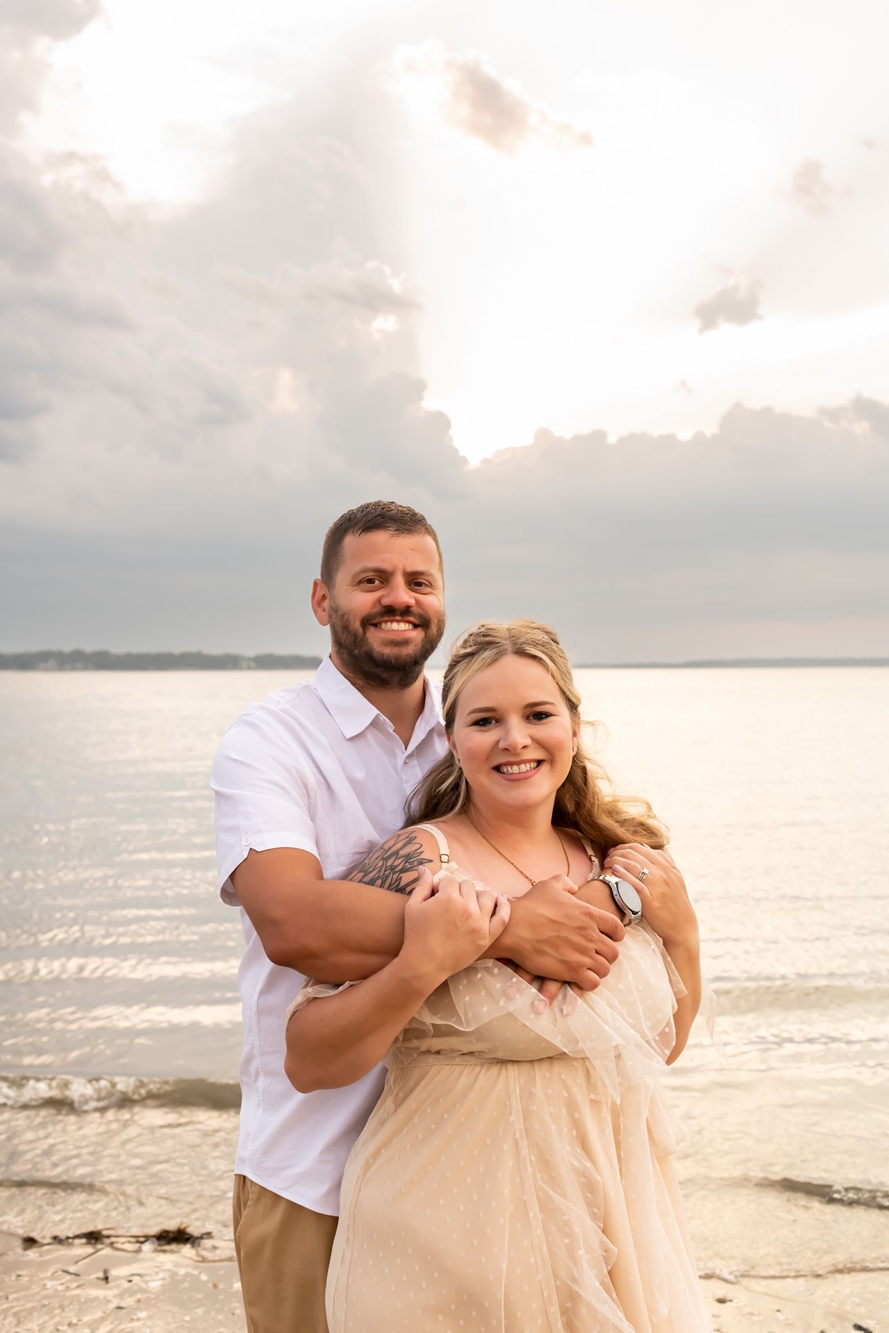 A couple embraces and laughs together on a beach at sunset with ocean waves in the background.