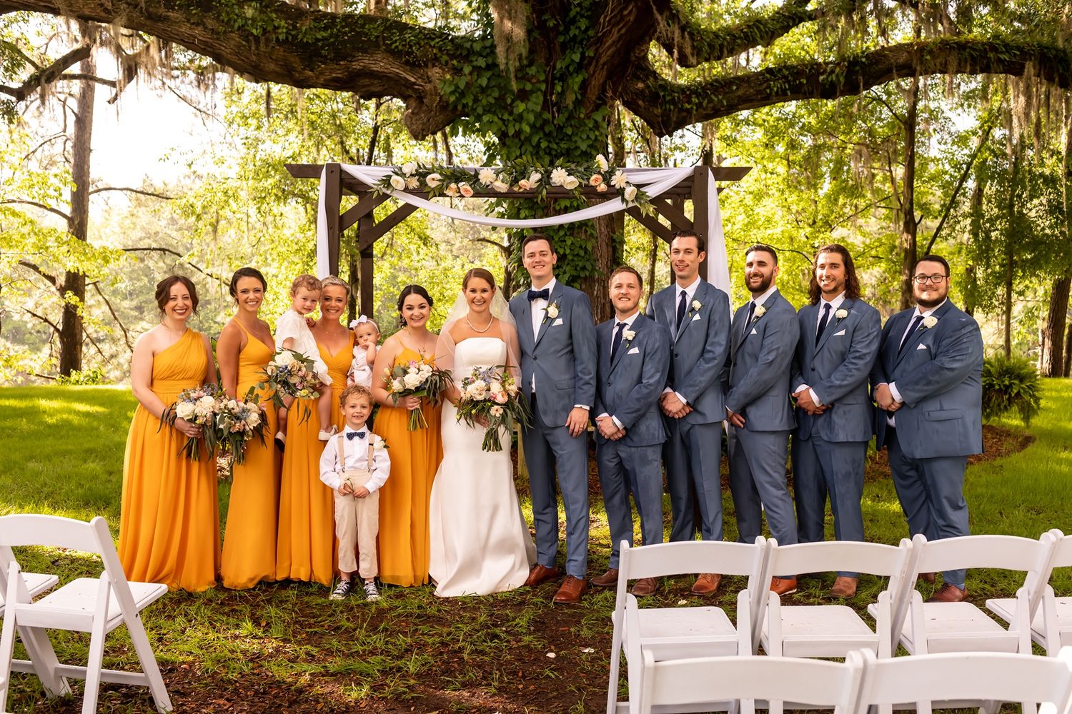 Wedding party members in orange dresses and navy suits pose together for outdoor group photos under shady trees.