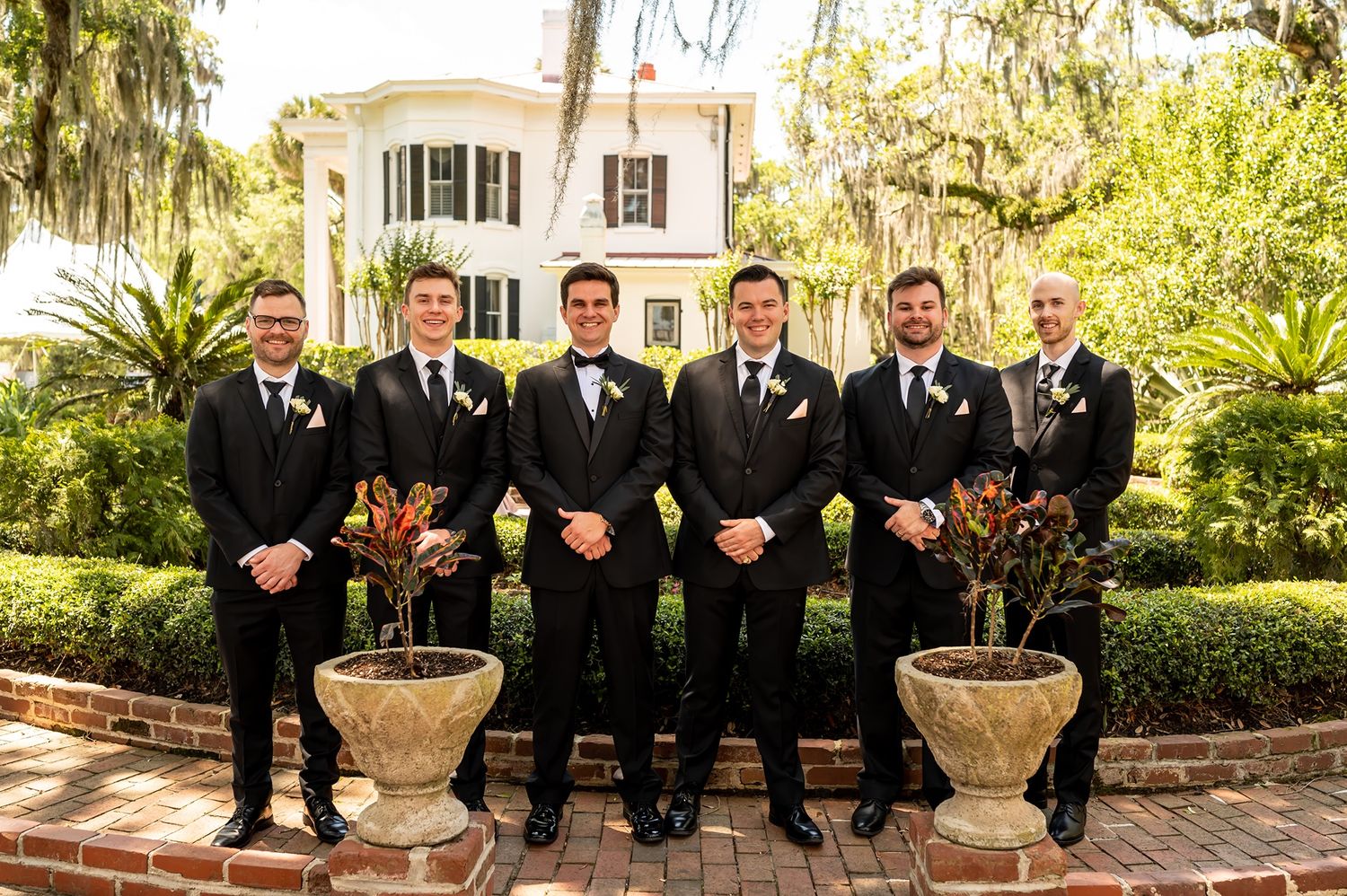 A group of groomsmen in black suits pose together on a paved patio at an elegant Southern mansion with lush gardens.