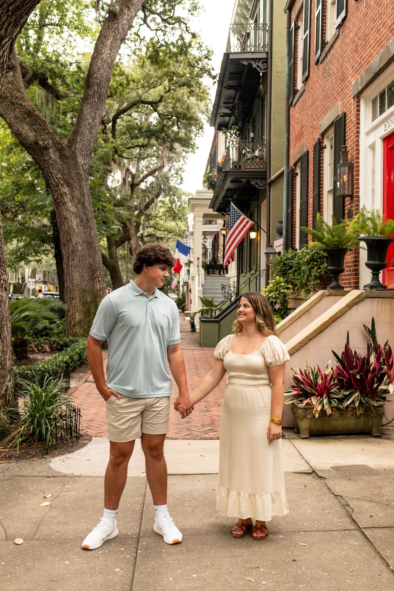 A couple holding hands on a historic brick sidewalk in Savannah Georgia with American flags and Spanish moss in trees above.