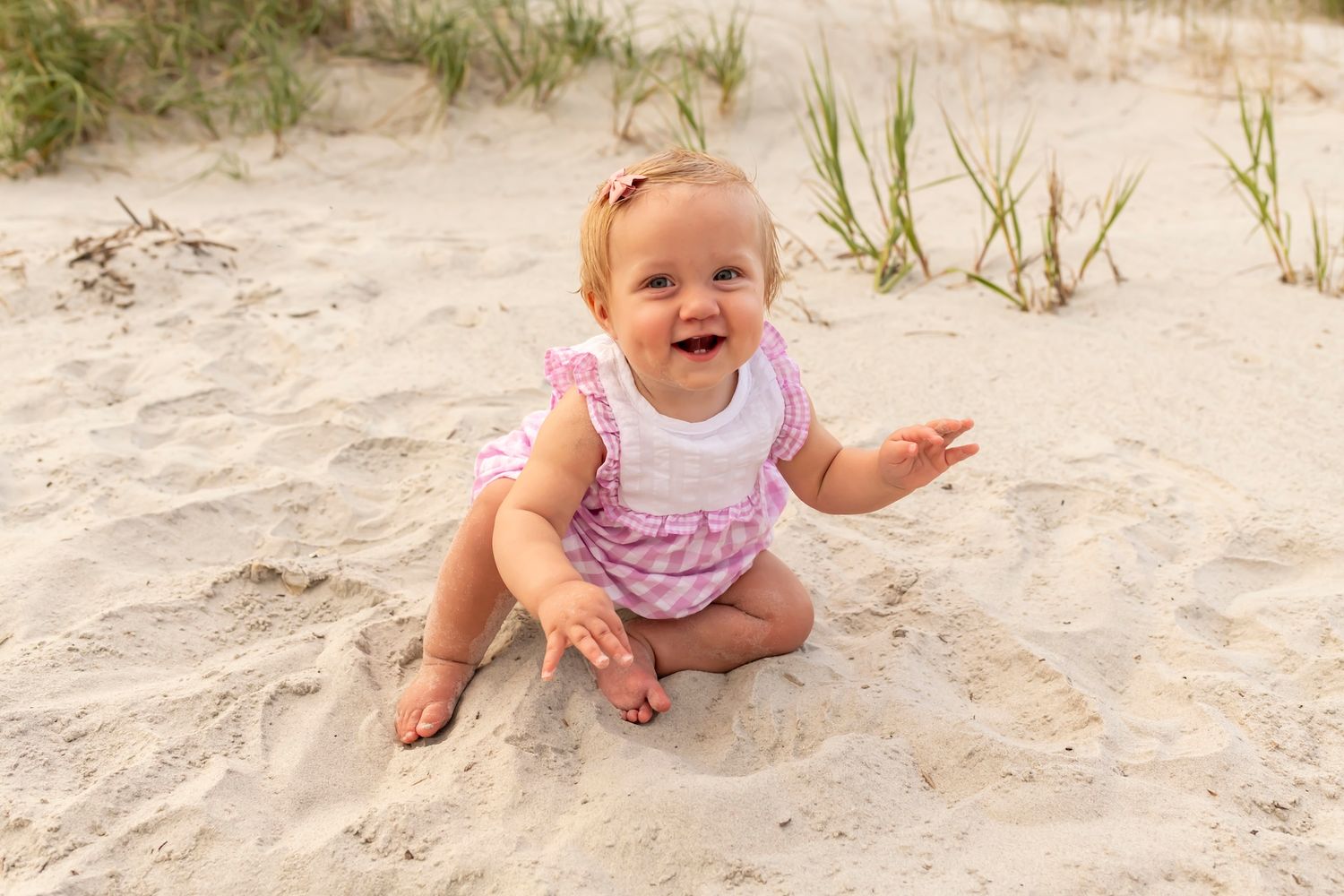 A joyful baby wearing pink and white clothing sits and plays in the sand on a sunny beach.
