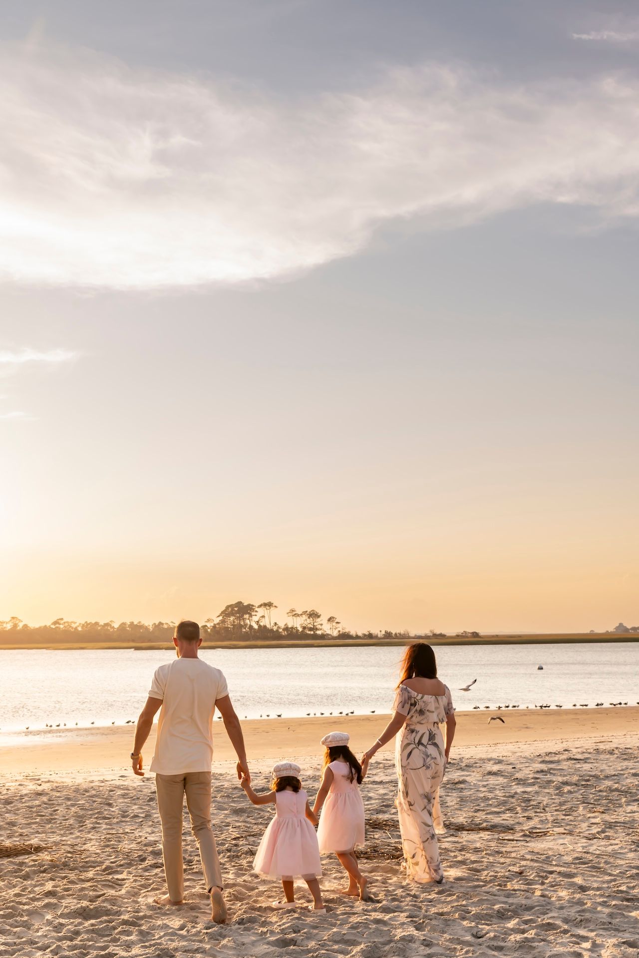 Family holding hands while walking on sandy beach at sunset with calm waters and trees in background.