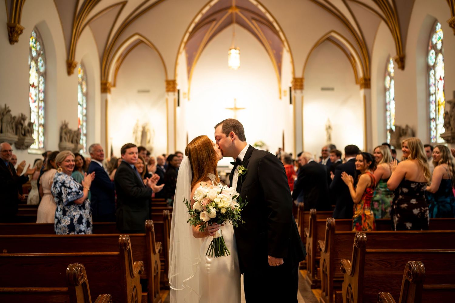 Romantic wedding kiss in a traditional church sanctuary with arched ceilings and stained glass windows.