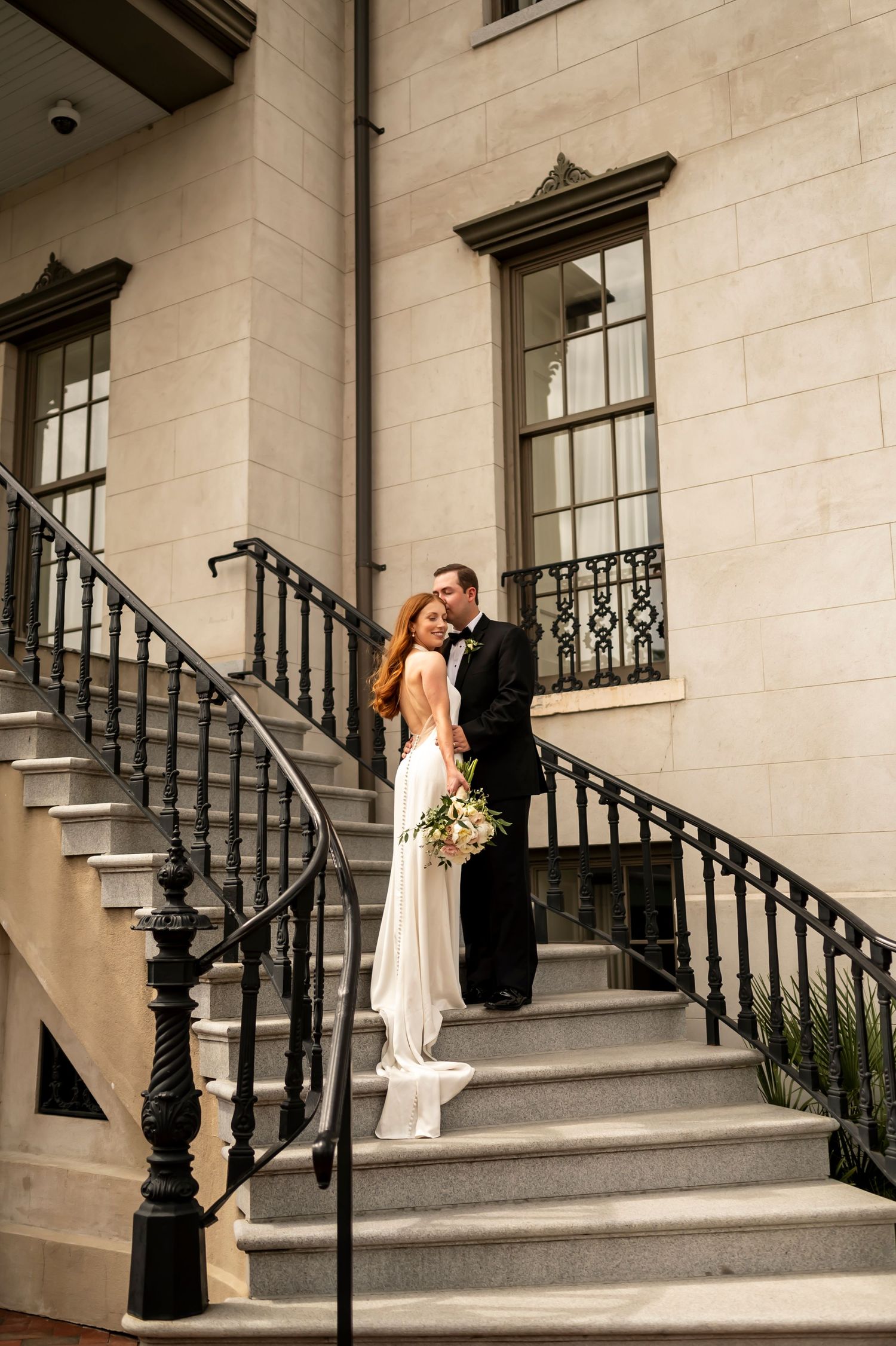 A romantic wedding couple stands on elegant stone steps with an ornate iron railing against a cream-colored building facade.