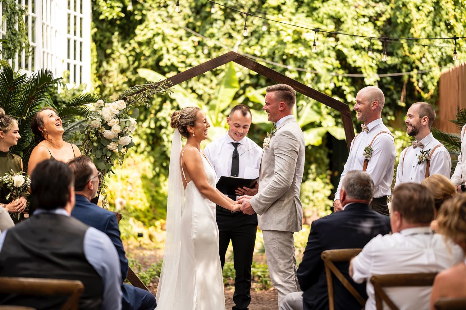A couple exchanges vows during an outdoor garden wedding ceremony surrounded by guests seated on wooden chairs.