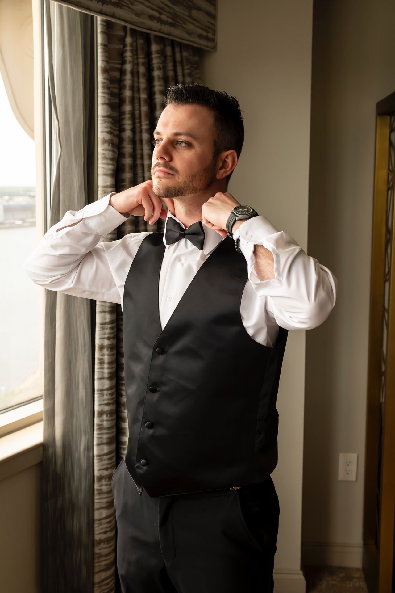 Formal dressed person adjusts black bow tie while standing by window in black vest and white dress shirt.
