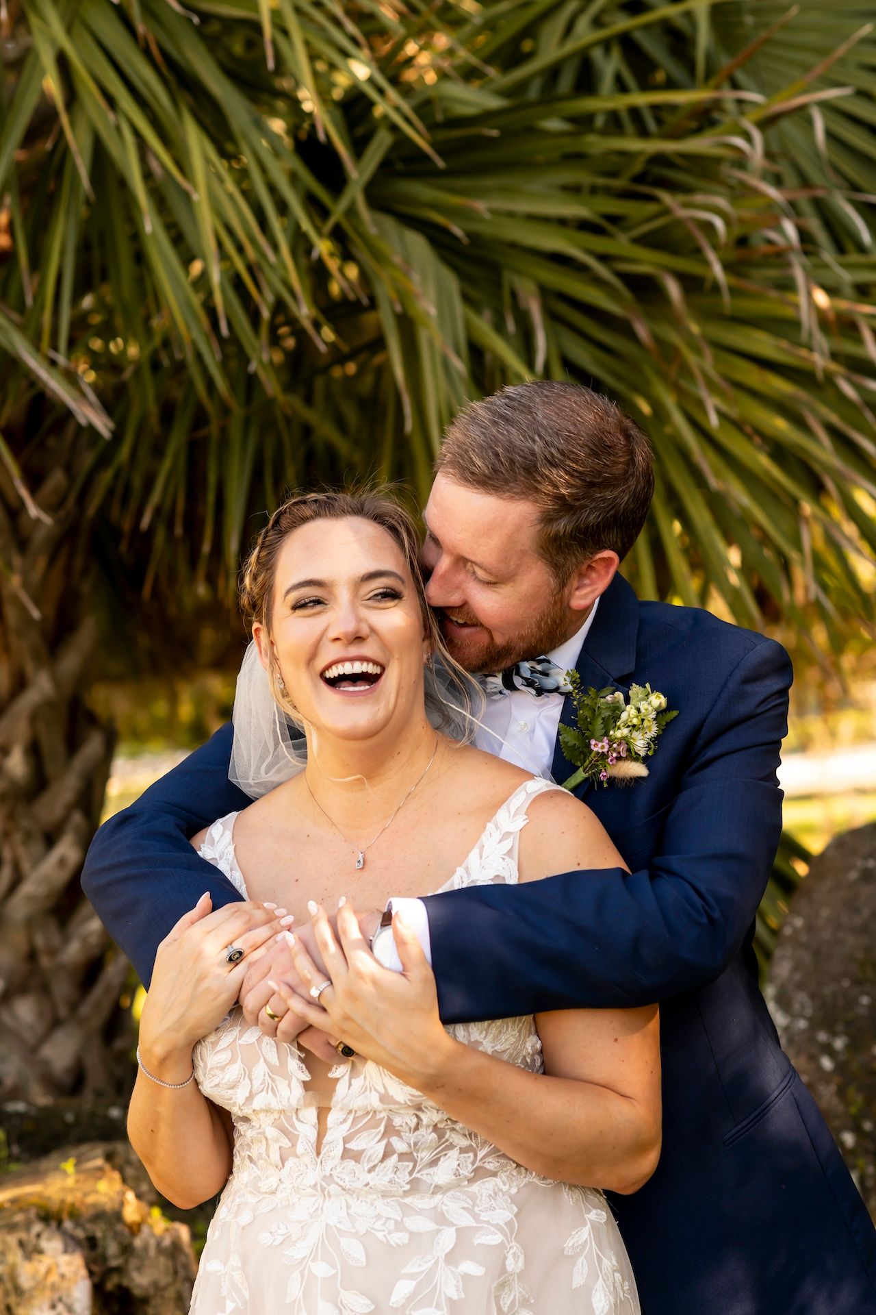 Newlyweds embrace joyfully against a backdrop of tropical palm fronds on their wedding day.