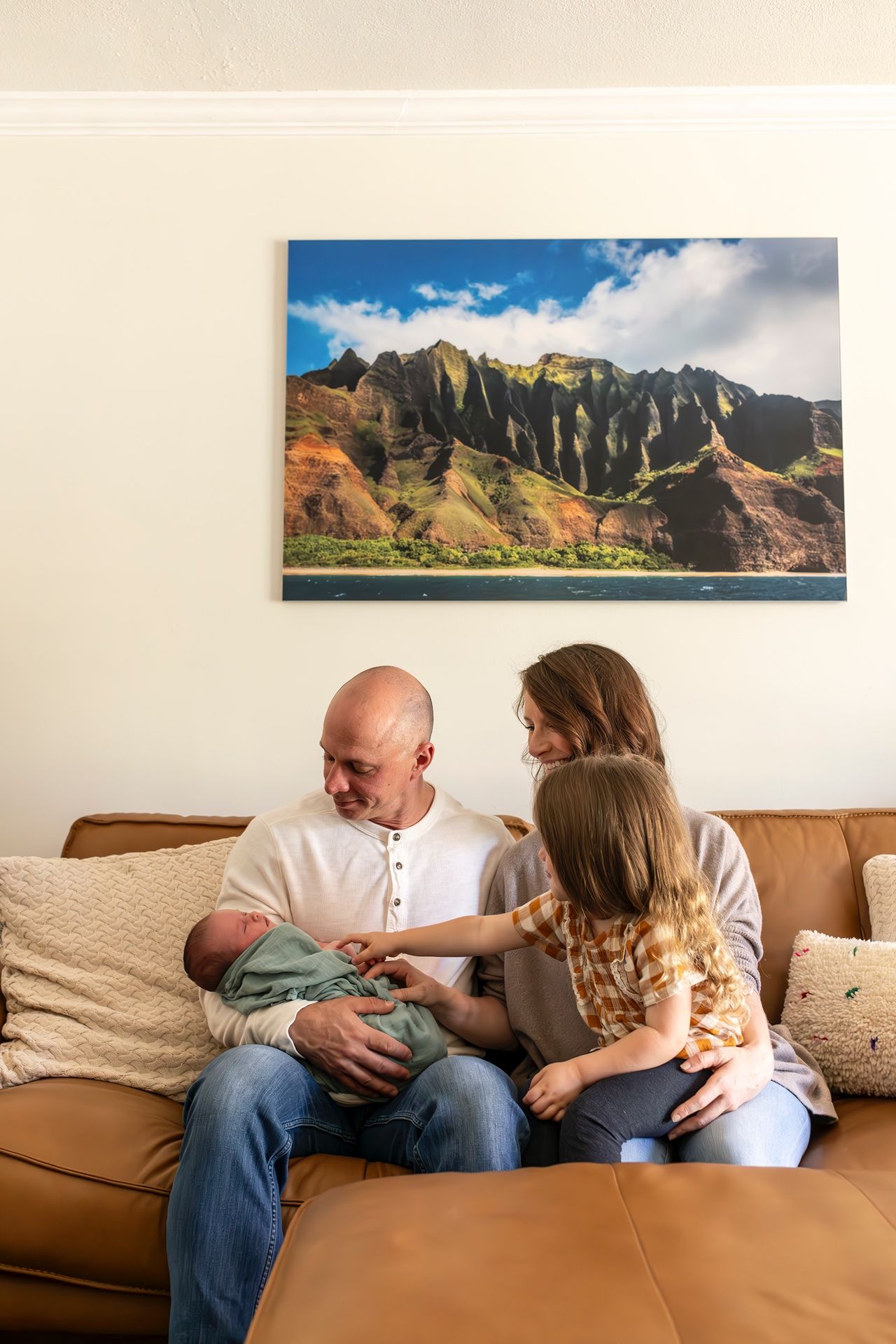 Family relaxing on brown leather couch with newborn baby under scenic mountain landscape art on wall.