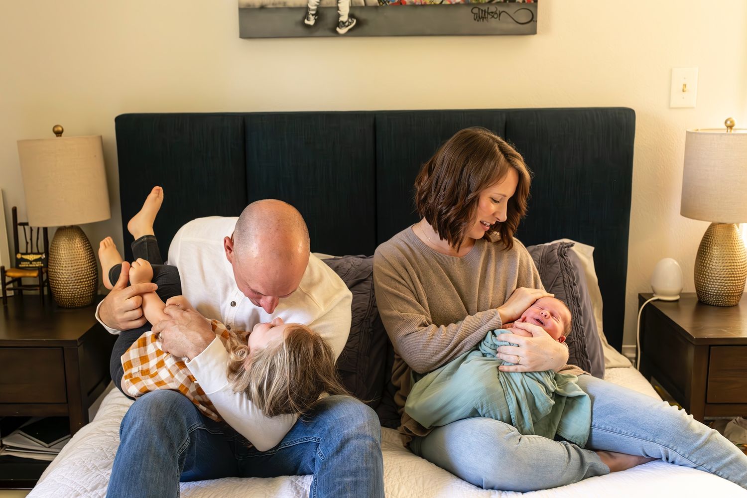 Two adults sitting on a bed comforting and bonding with babies in a cozy bedroom setting with lamps and decor.
