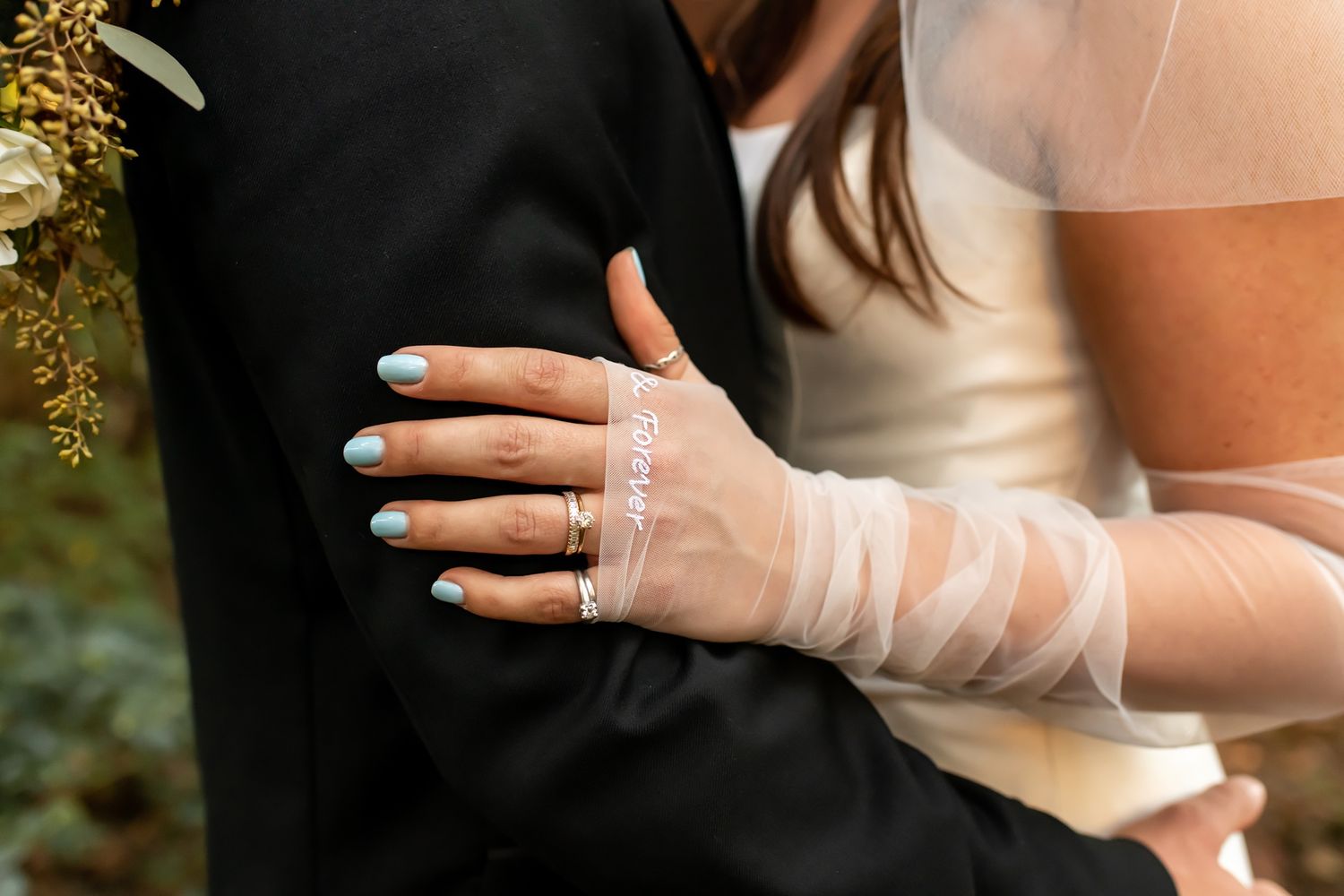 A close-up of a bride's hands wearing sheer white gloves resting on a black suit jacket.