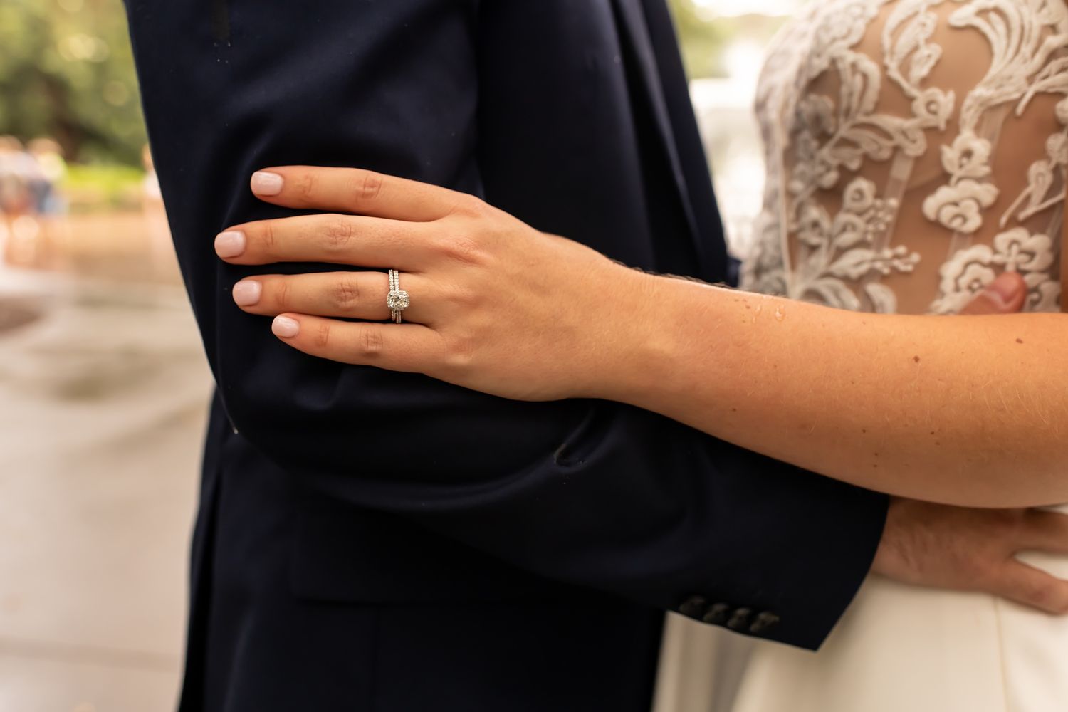 A bride's hand rests on a groom's navy suit sleeve, showing off a sparkling wedding ring in natural daylight.