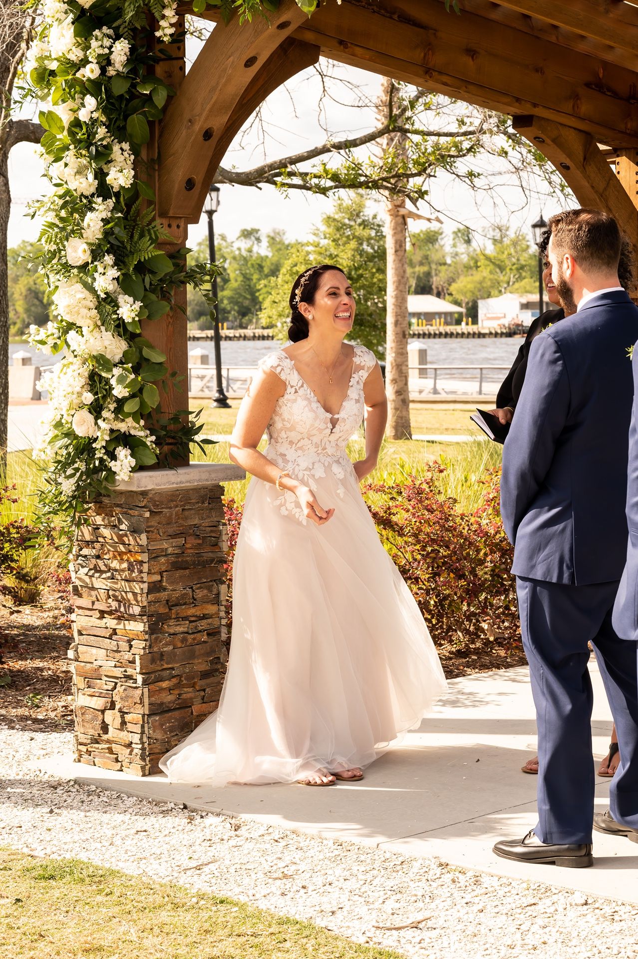 Couple exchanges vows under a wooden arch decorated with white flowers during sunny outdoor wedding ceremony.