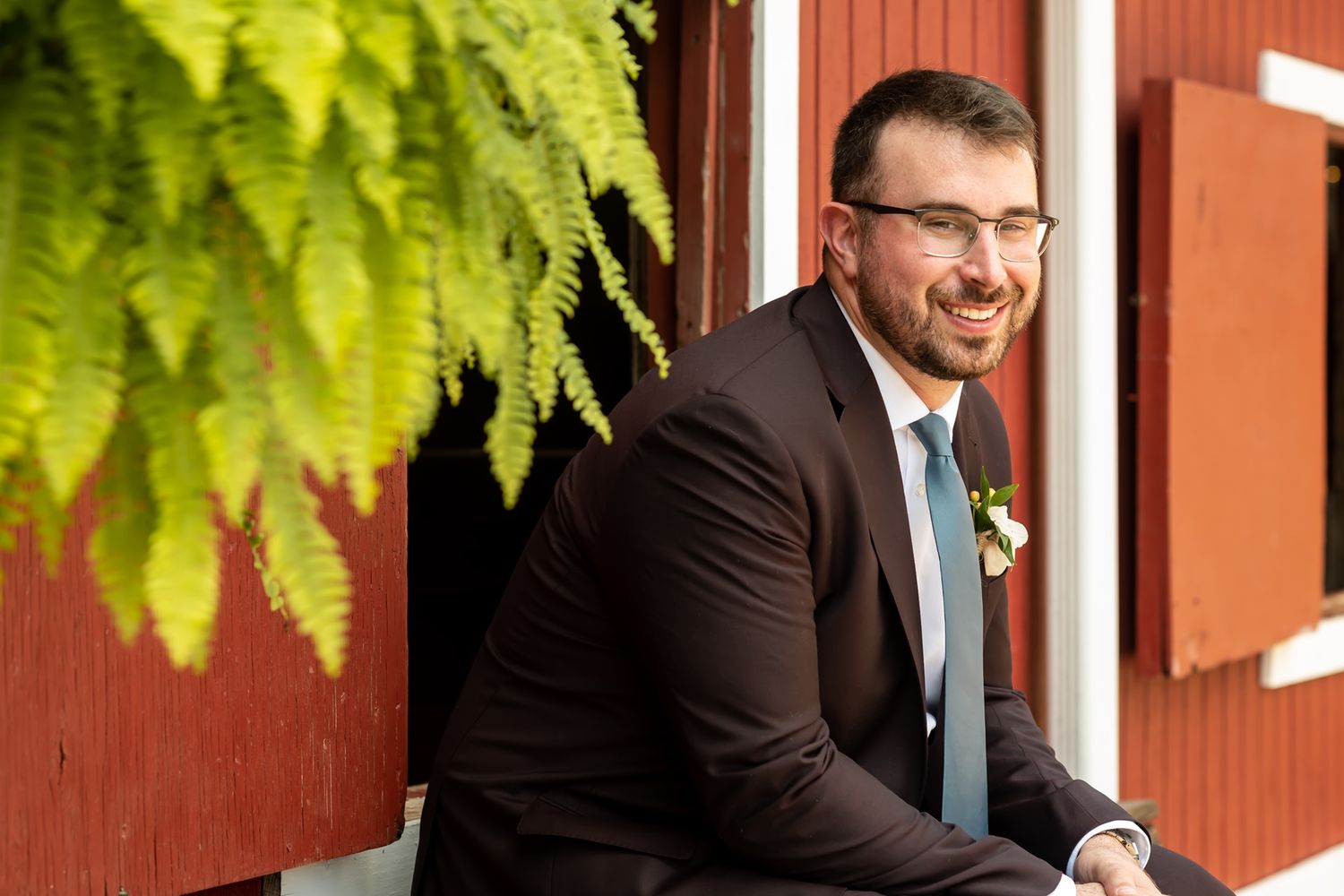 Groom in a navy suit smiles while sitting on wooden steps with greenery and red barn walls in the background.