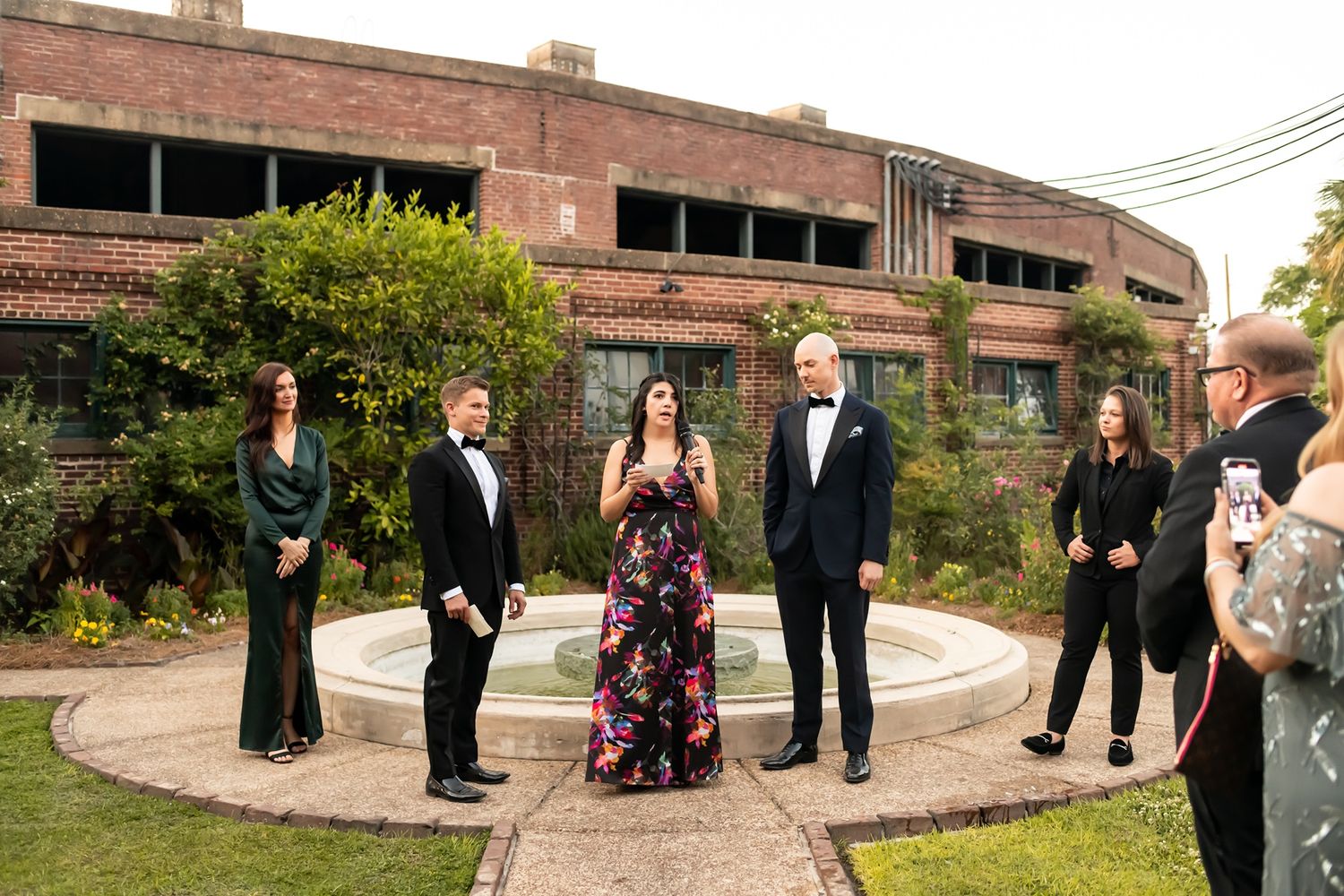 Formal group in evening attire poses in front of rustic brick building with landscaped garden courtyard at sunset.