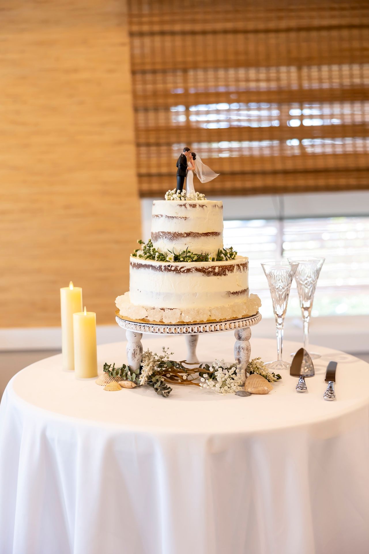 Three-tiered white wedding cake with greenery accents and bride and groom topper displayed on elegant table setting.