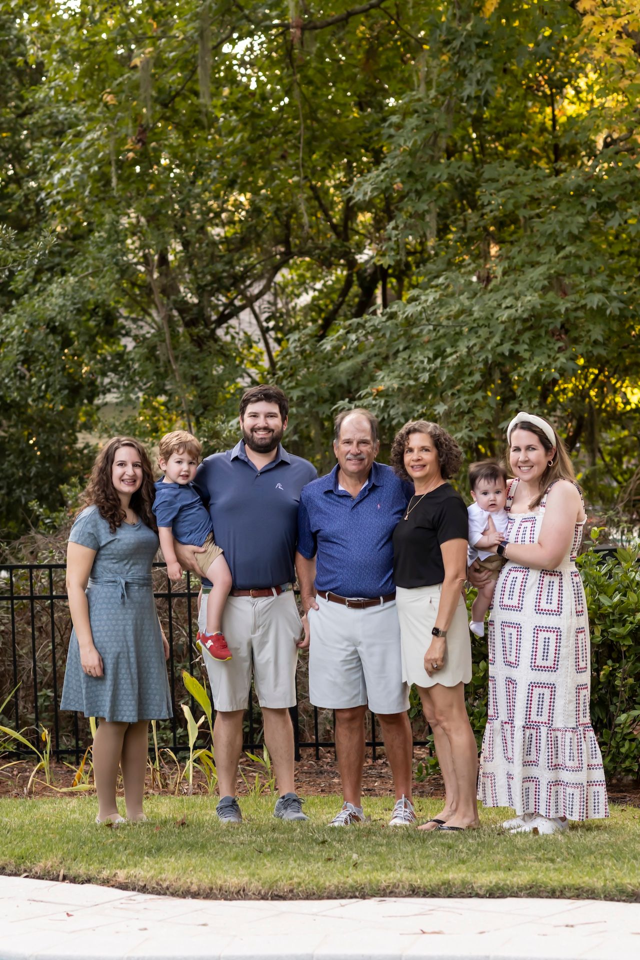 A multi-generational family group poses together outdoors on a lawn with green trees and foliage in the background.