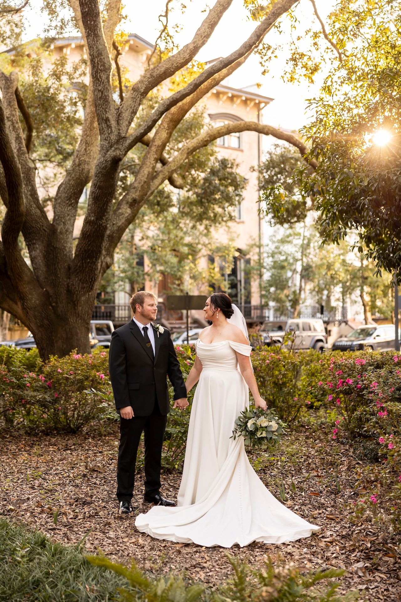 A couple in wedding attire stands in a garden with blooming flowers under a large tree during golden hour sunset.
