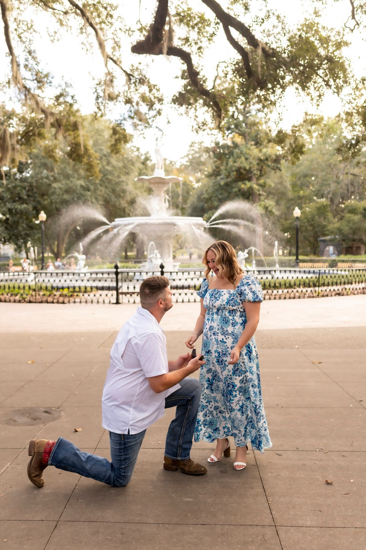 A romantic proposal scene unfolds on a concrete plaza beneath sunlit tree branches.