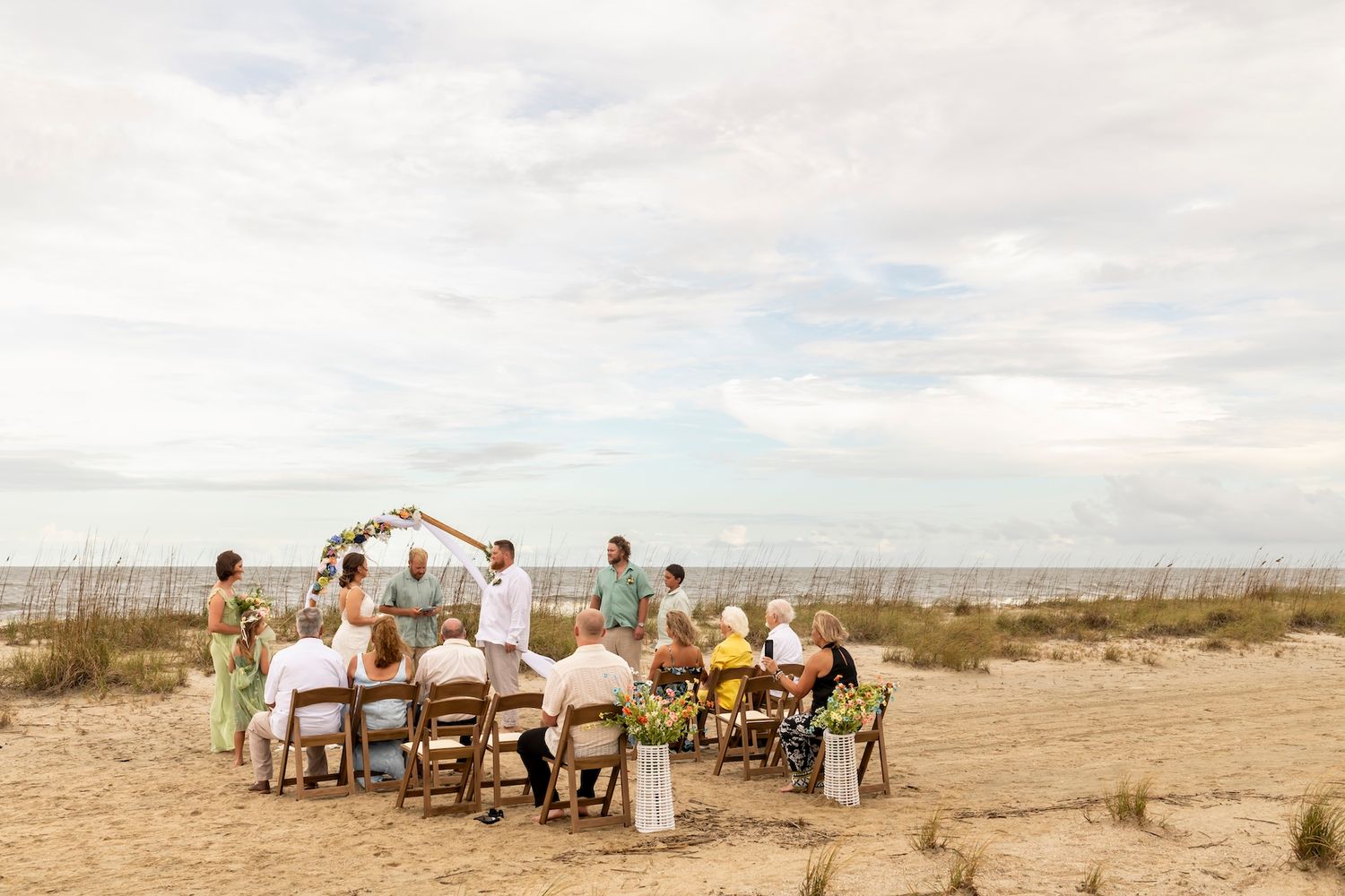 A small intimate wedding ceremony takes place on a sandy beach with guests seated in white chairs under cloudy skies.
