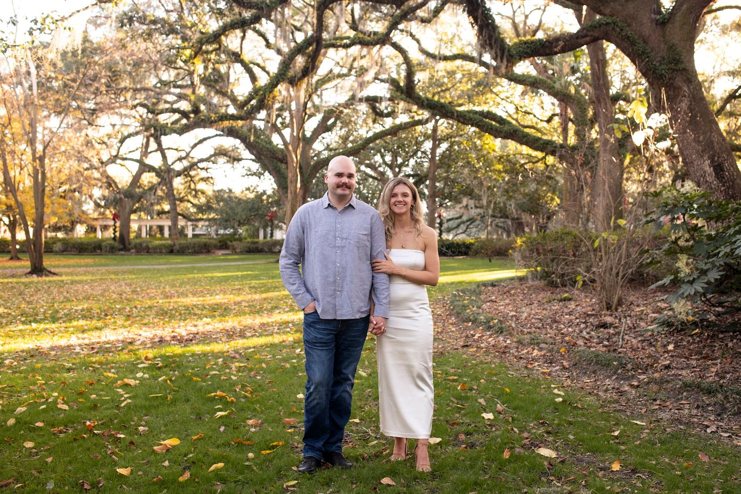 A couple stands together outdoors under majestic oak trees in a serene park setting during autumn.