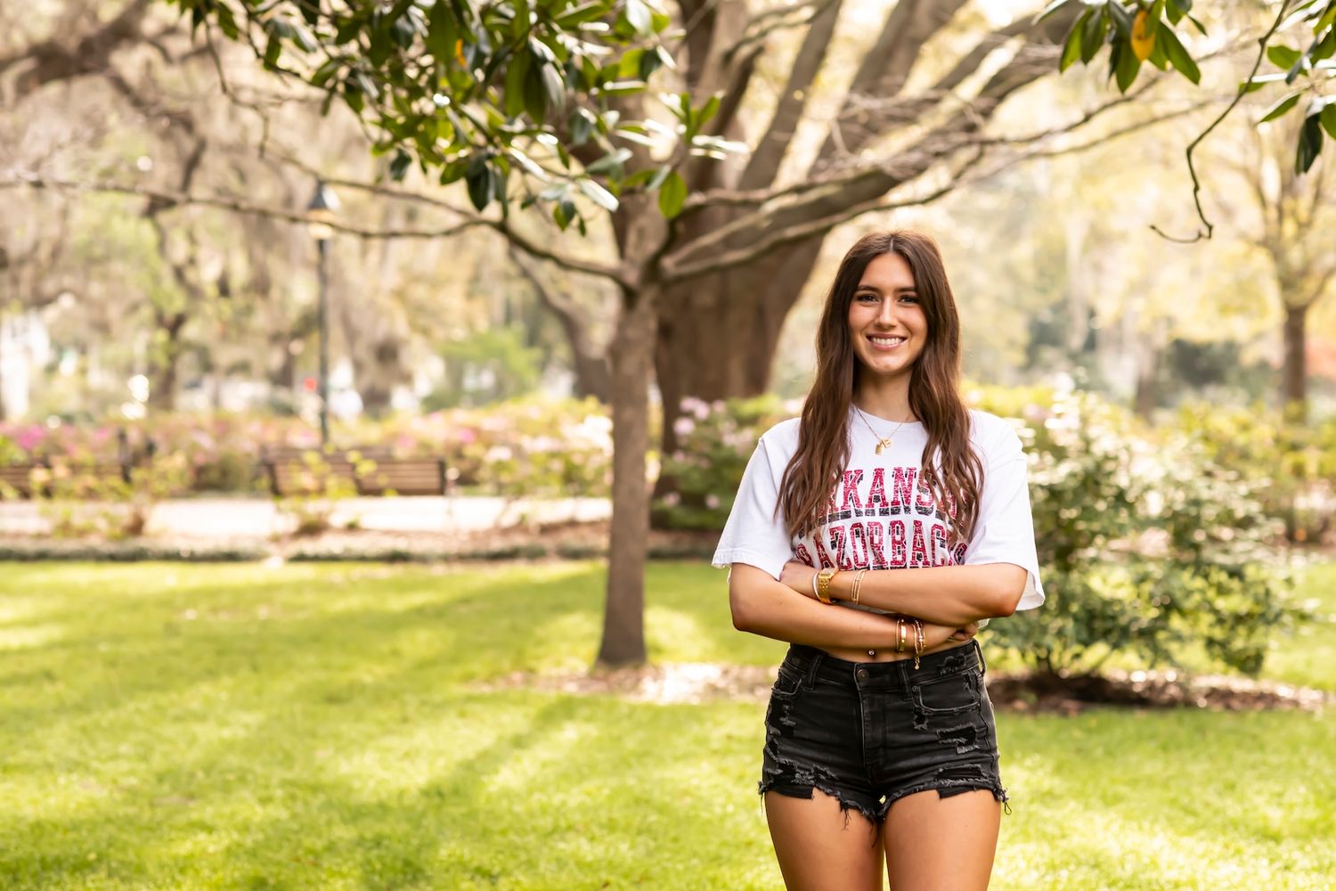 A person wearing a university t-shirt poses outdoors on a sunny day in a park-like setting.