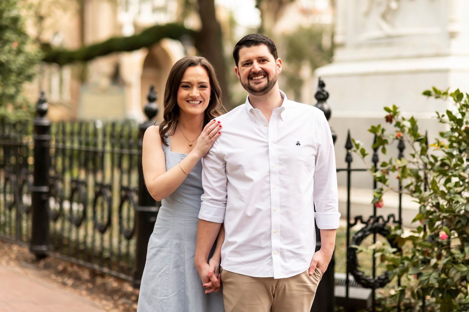 A couple smiles while holding hands on a sunny day beside an ornate iron fence and greenery.