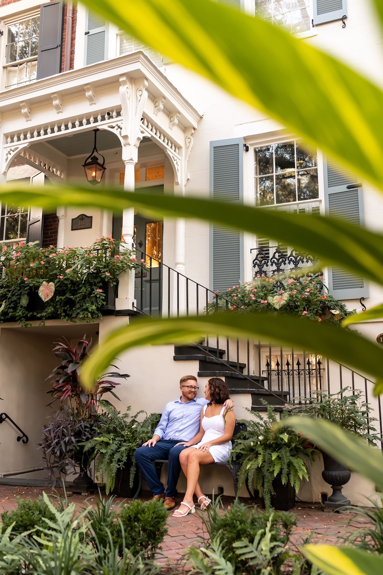 Two people sit together on steps of a charming Victorian house with ornate details and lush tropical landscaping.