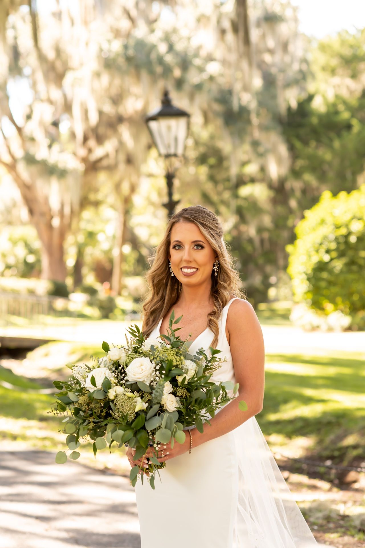 A bride in a white gown holds a lush green and white floral bouquet while standing on a sunlit garden path.